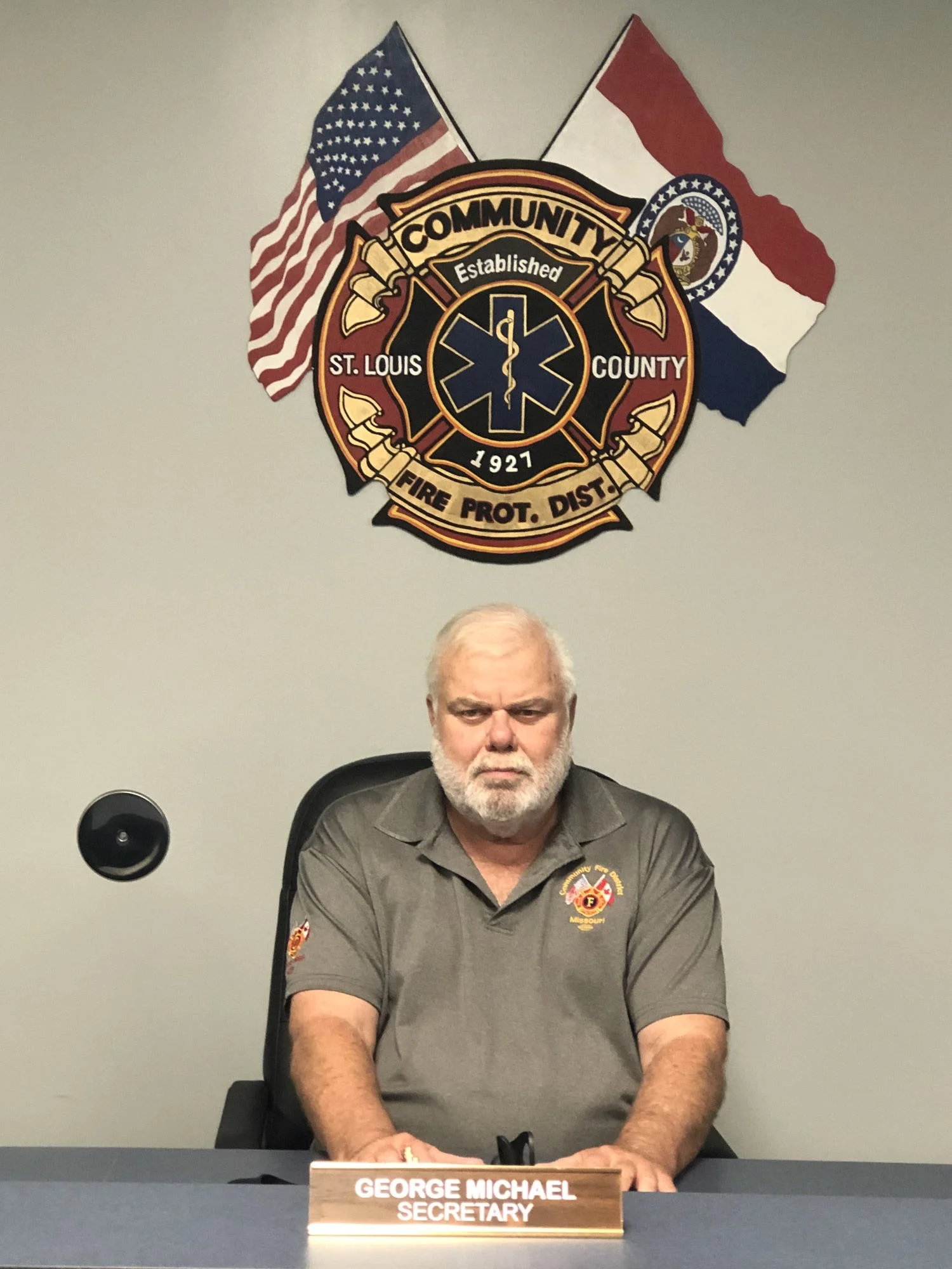 A man with a beard and gray hair sitting at a desk with a nameplate reading "George Michael, Secretary." He is wearing a gray shirt with fire department insignia and sitting in front of a wall with fire department and American flags, and a fire department badge emblem.