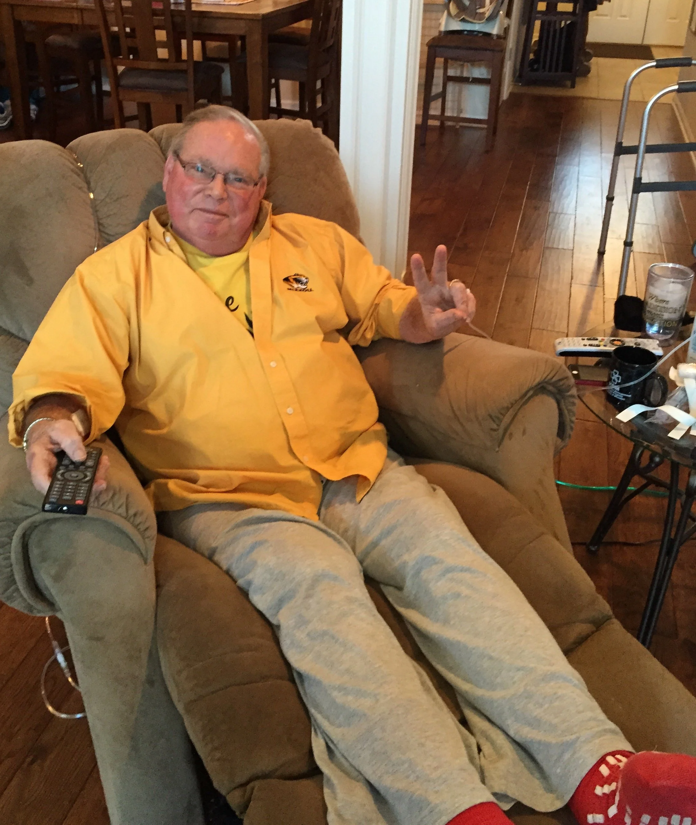 A man with glasses and wearing a yellow shirt is sitting on a beige couch, making a peace sign with his right hand. His legs are extended on the couch, and he is wearing red shoes. The background shows a wooden dining table, chairs, a walker, a glass of water, television remote, and some miscellaneous items on a small table.