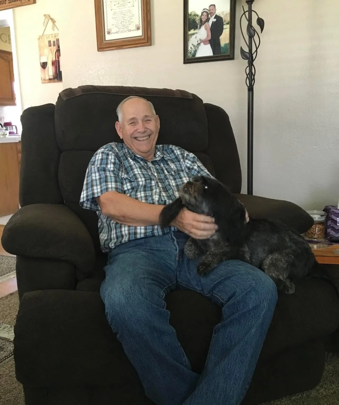 A smiling man sitting on a black armchair, holding a black and brown dog in his lap. The man is wearing a plaid shirt and blue jeans, and the dog is looking up at him. Behind them are framed pictures of a wedding and a man and woman on the wall, and a tall floor lamp is nearby.