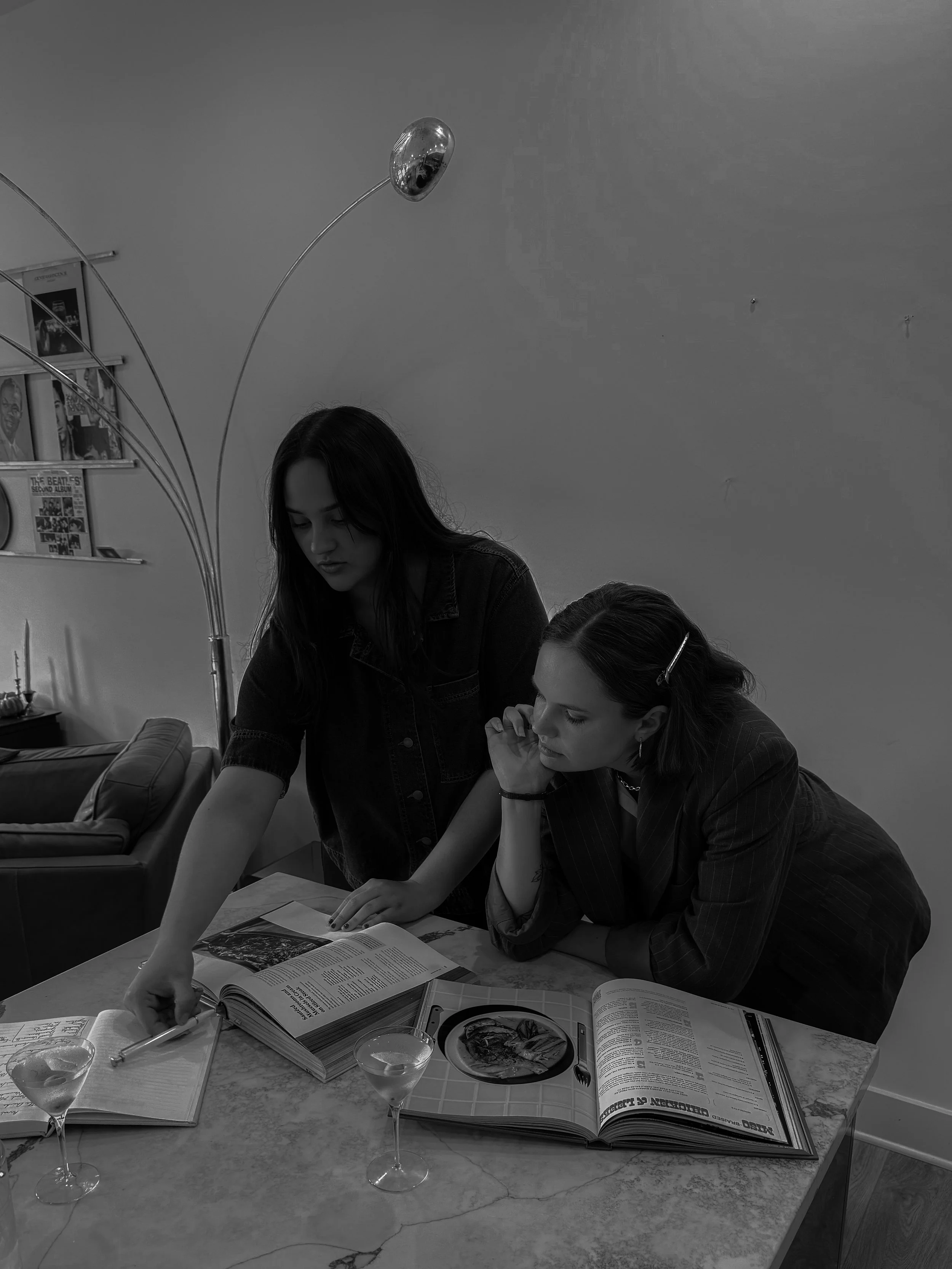 Two women sitting at a table with open books, one pointing at a page, with drinks and plates on the table, in a room with a bookshelf and a modern lamp.