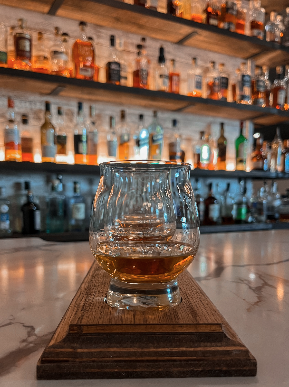 A glass of whiskey on a wooden tray on a bar counter with shelves of liquor bottles in the background.