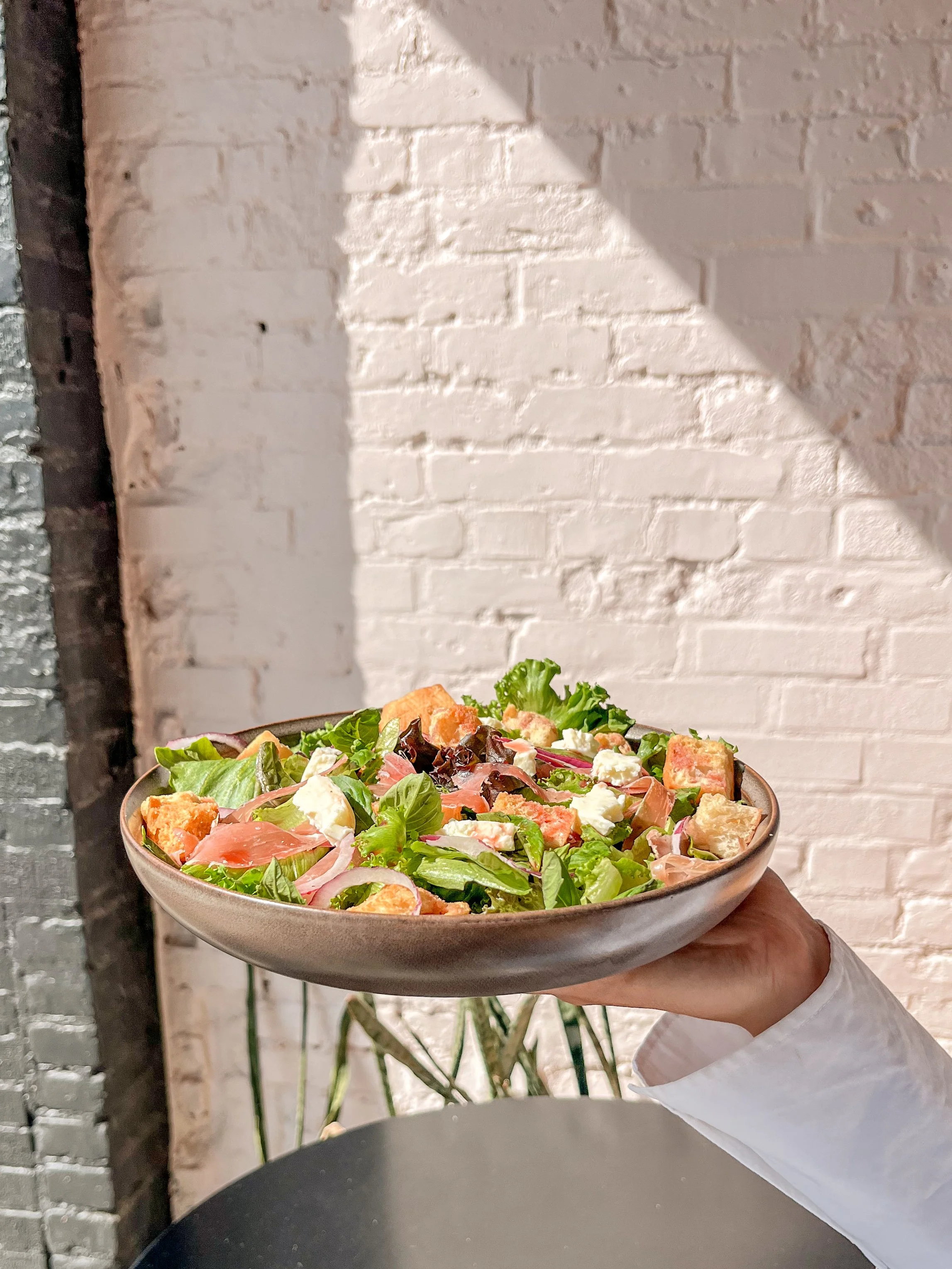 A person holding a bowl of salad with leafy greens, tomatoes, and cheese, against a white brick wall background.