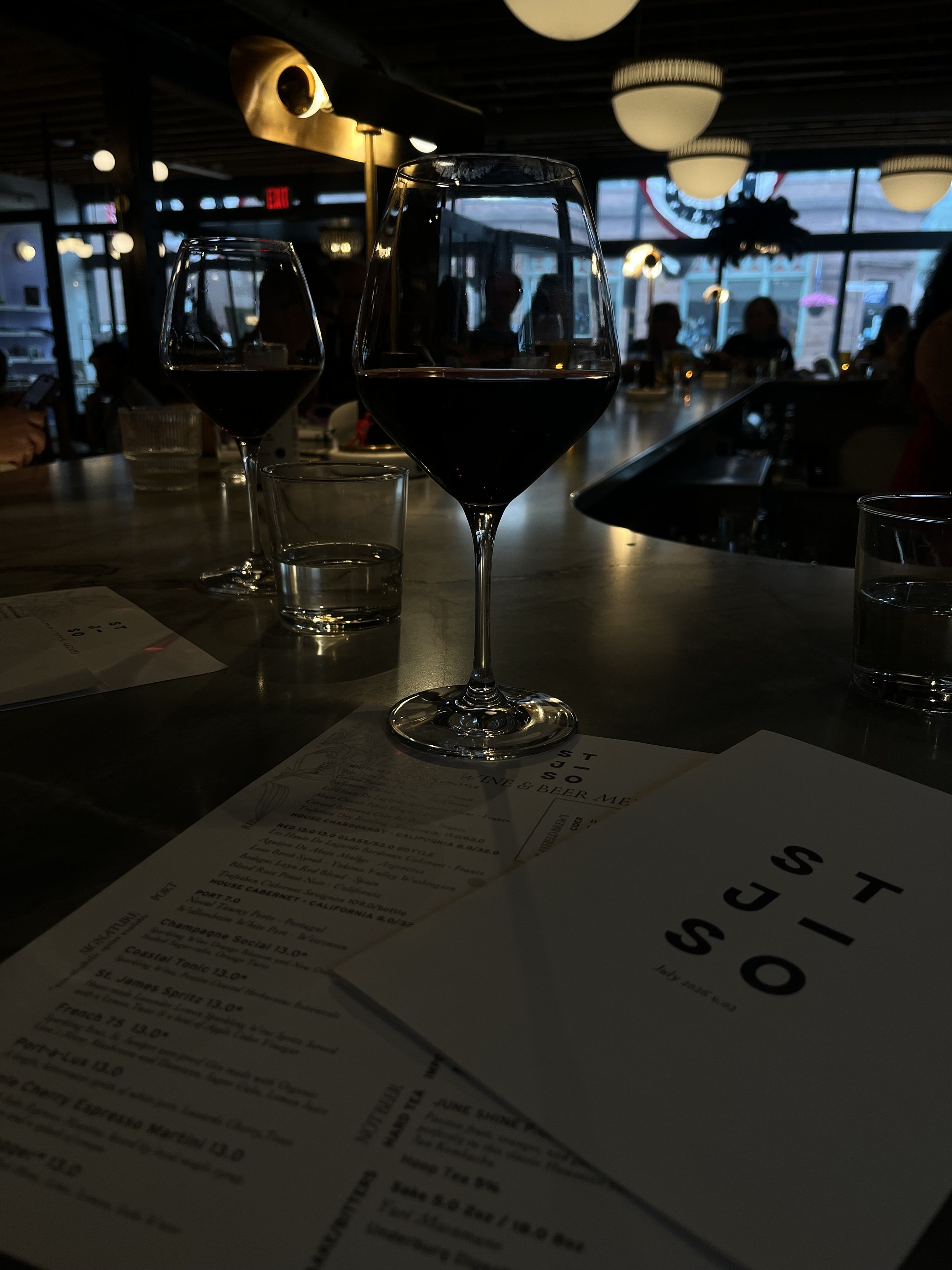 Two glasses of red wine on a restaurant table with a menu, in a dimly lit restaurant with hanging light fixtures and large windows in the background.