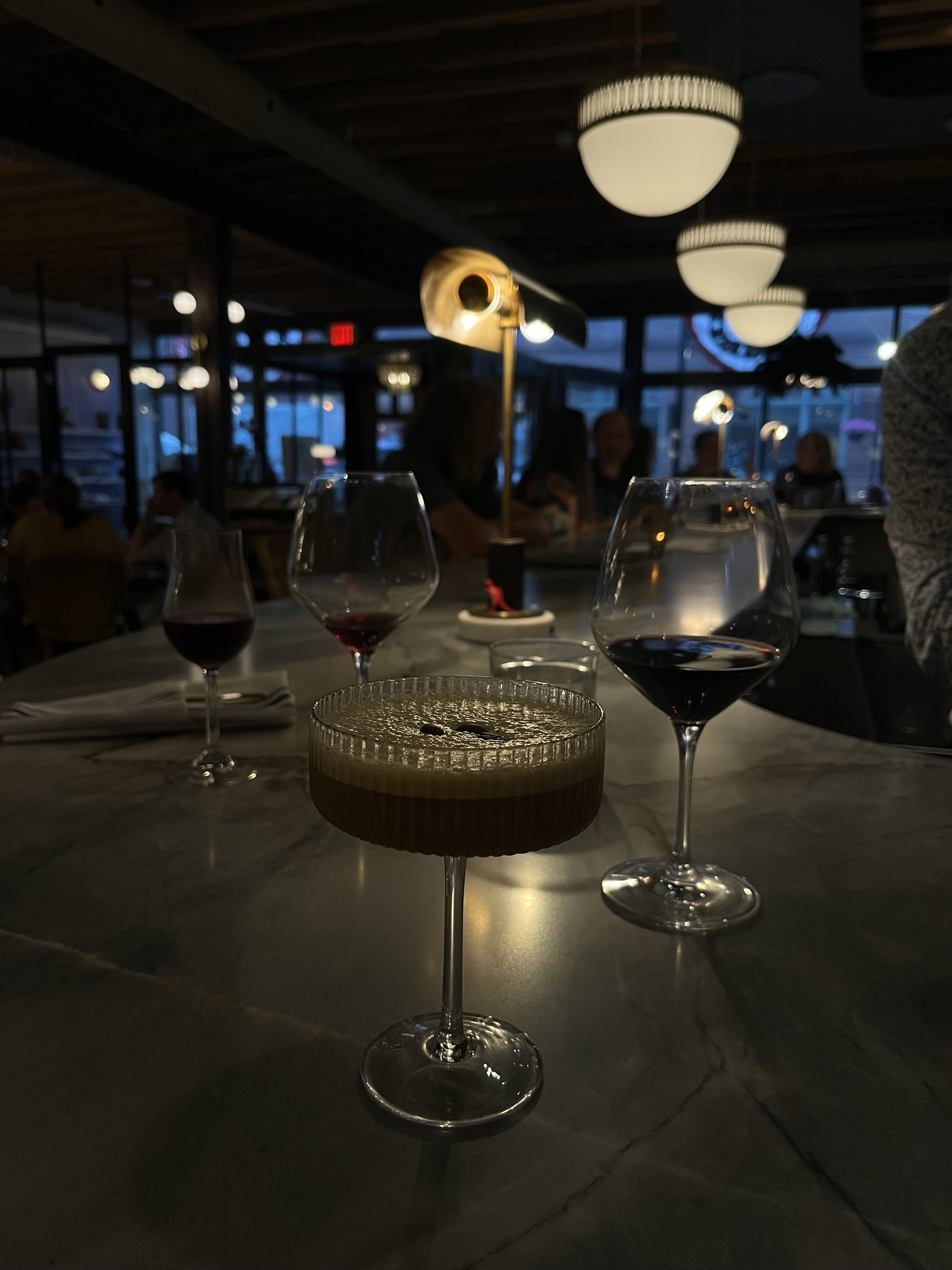 A restaurant scene in the evening with wine glasses, a lit candle, and people sitting at tables. The dim lighting and warm ambiance are visible, with modern ceiling lights and large windows in the background.