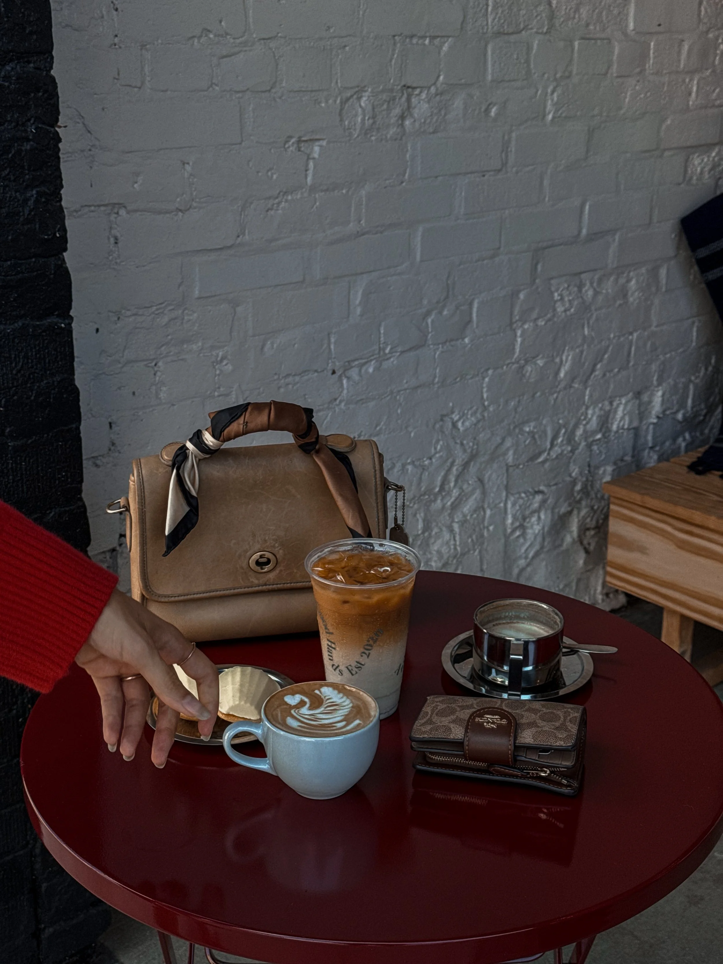 A red round table with two cups of coffee, one with latte art, a glass of iced coffee, a small jar of water, a wrapped chocolate, and a small leather wallet. A person's hand is reaching for the cup of latte.