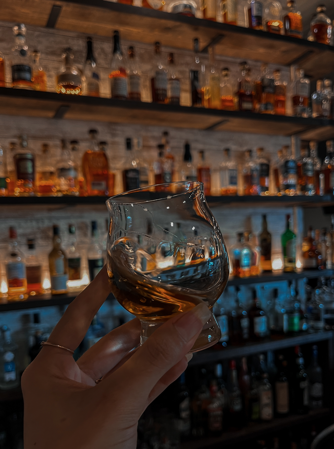 A hand holding a glass of whiskey in front of a shelf filled with various bottles of liquor.