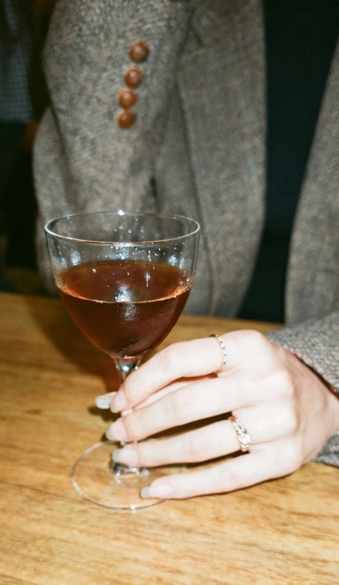A person holding a glass of red wine with their left hand, wearing rings, and dressed in a grey blazer with brown button details on the sleeve.