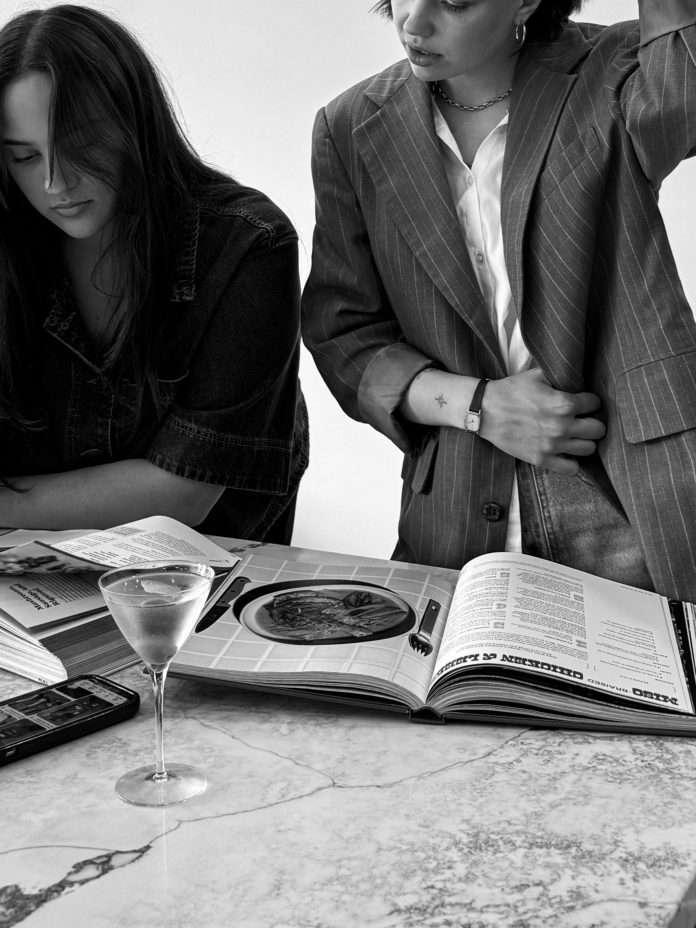 A person with long dark hair and a denim jacket leaning over a table, a woman with short dark hair and striped blazer standing next to her, with open magazines and a glass of water on the marble table.