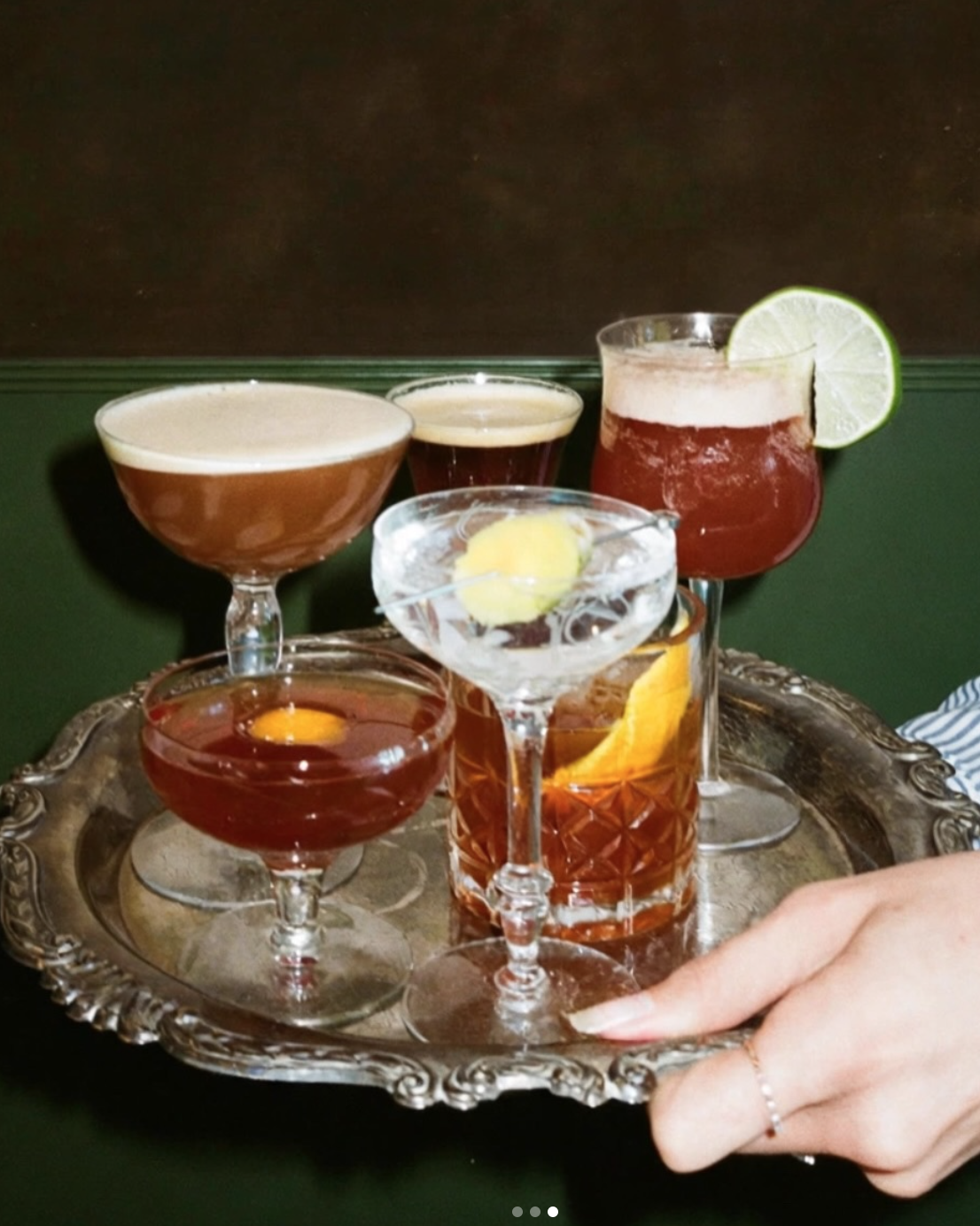 A silver tray holding six different cocktails with varying colors, garnished with lime slices and lemon peel, against a dark background.