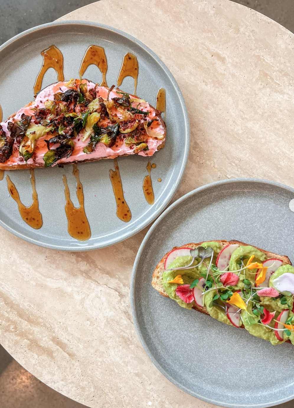 Two plates with toasted bread topped with avocado and colorful edible flowers and microgreens, one has radish slices, the other has a pink spread with herbs and balsamic glaze.