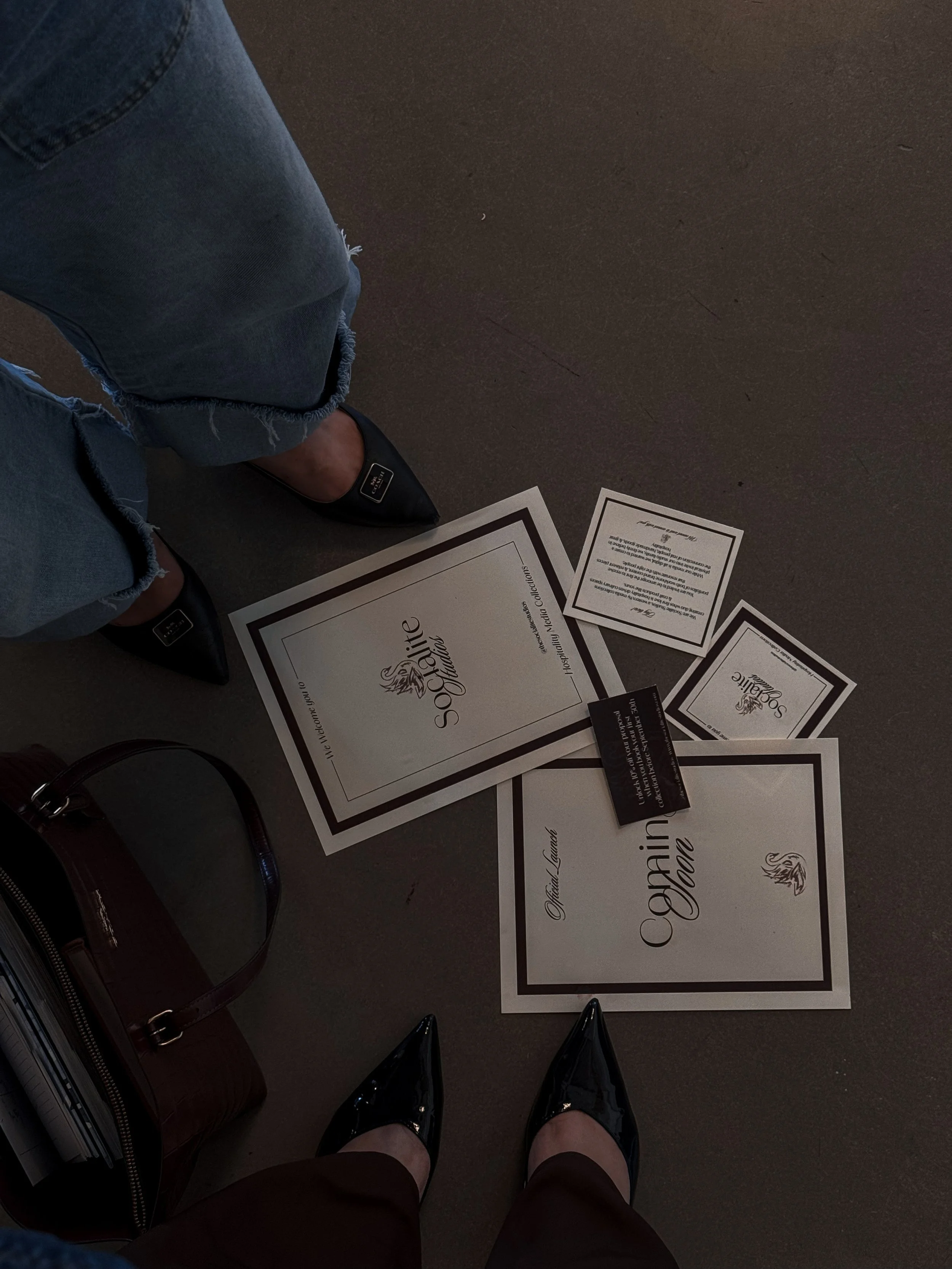 A person's legs wearing black high heels and ripped jeans are seen from above, standing on a floor among various documents and a black handbag.