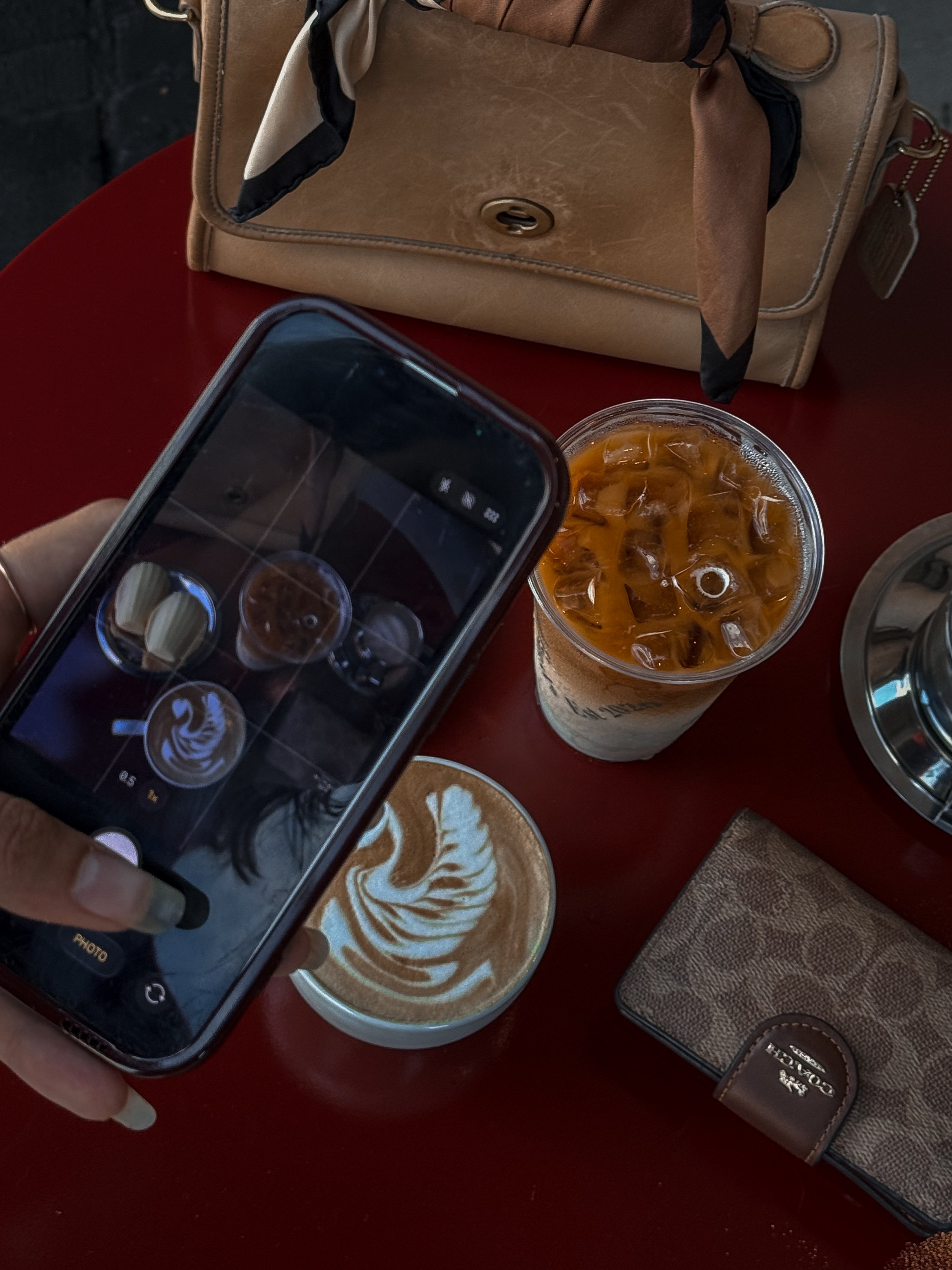 A person's hand holding a smartphone taking a photo of a latte art coffee and an iced coffee drink on a red table, with a tan purse and a leather wallet nearby.