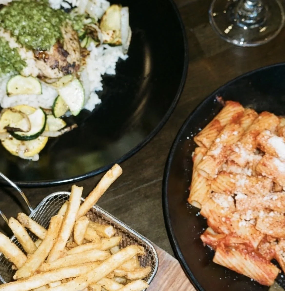 Black bowls containing grilled zucchini and chicken, a plate of cooked rigatoni pasta with tomato sauce and grated cheese, and a basket of French fries.