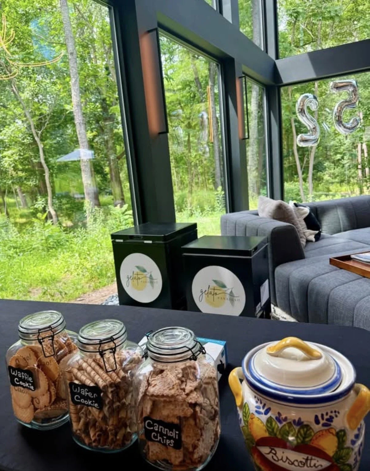 Indoor seating area with large glass windows revealing a green forest outside. On the table in the foreground are three jars of cookies labeled 'Waffle Cookie,' 'Wafer Cookie,' and 'Cannoli Chips,' and a colorful ceramic jar labeled 'Biscotti.'