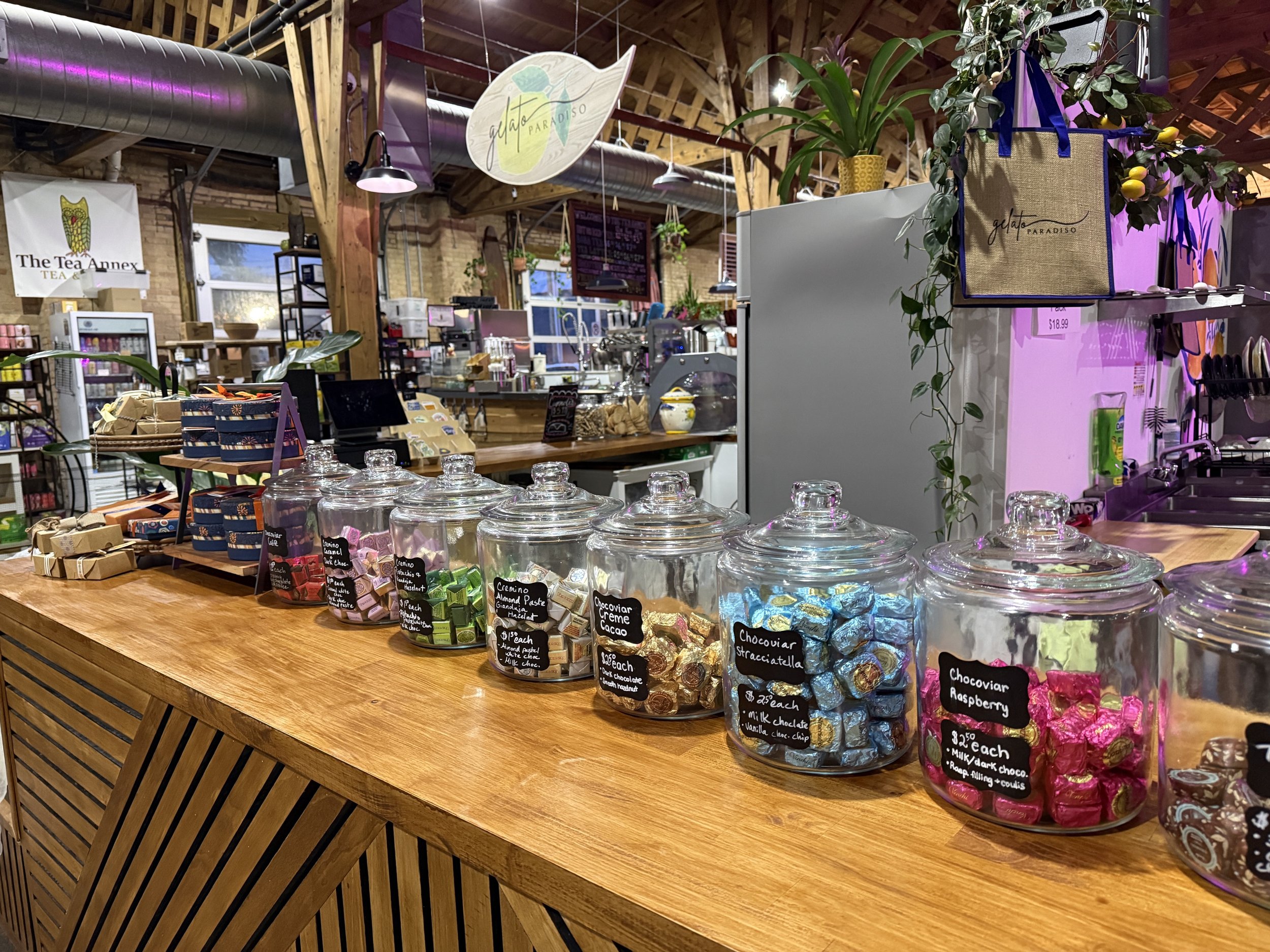 Inside a shop with a wooden counter displaying glass jars filled with wrapped chocolates. There are signs with flavor descriptions and prices. The background has a rustic ceiling with exposed wooden beams and industrial ventilation ducts, with plants and shelves.
