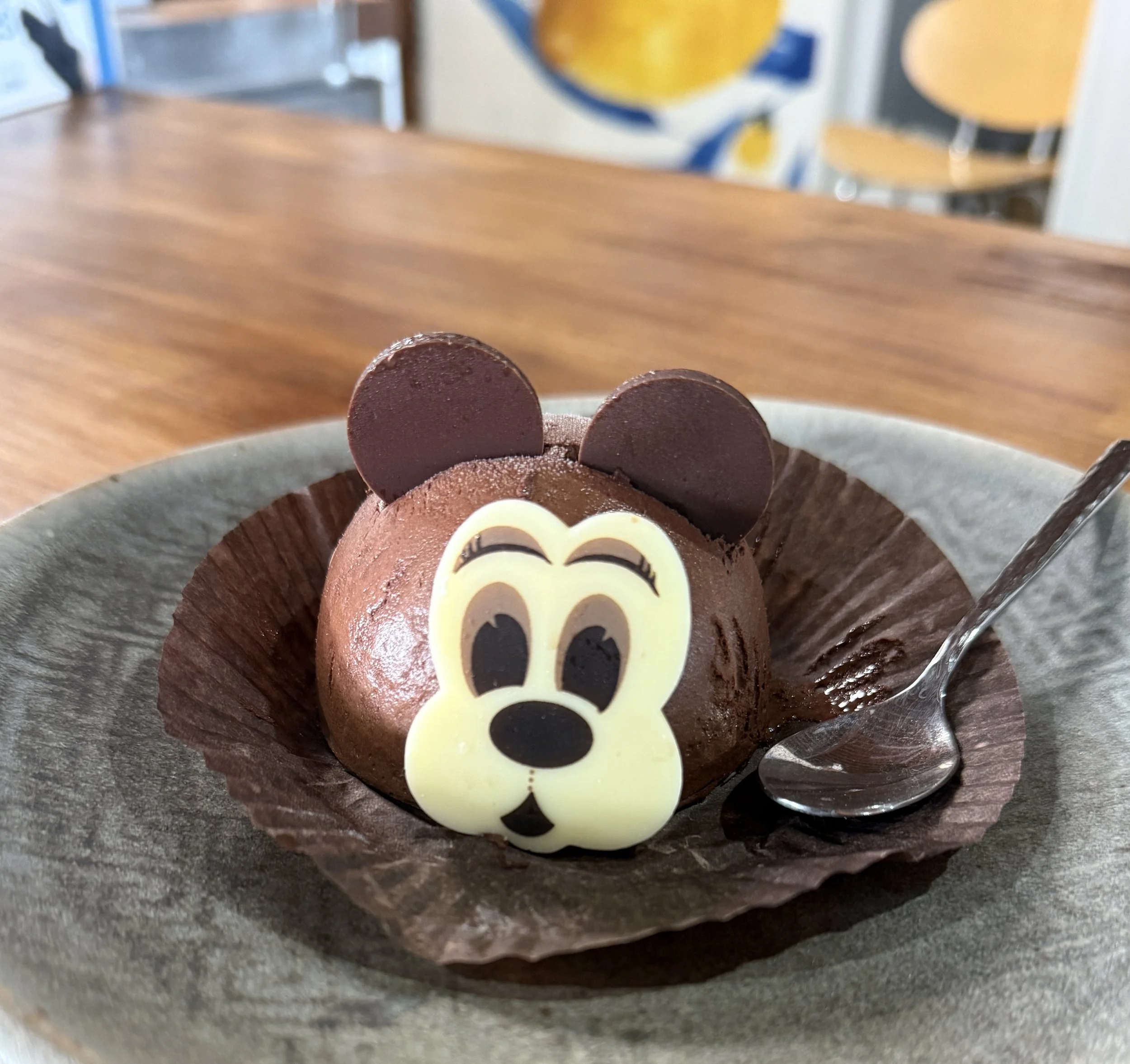 Close-up of a chocolate gelato bear with decorated eyes, nose, and a big smile, topped with white chocolate eyes and chocolate ears, on a plate.