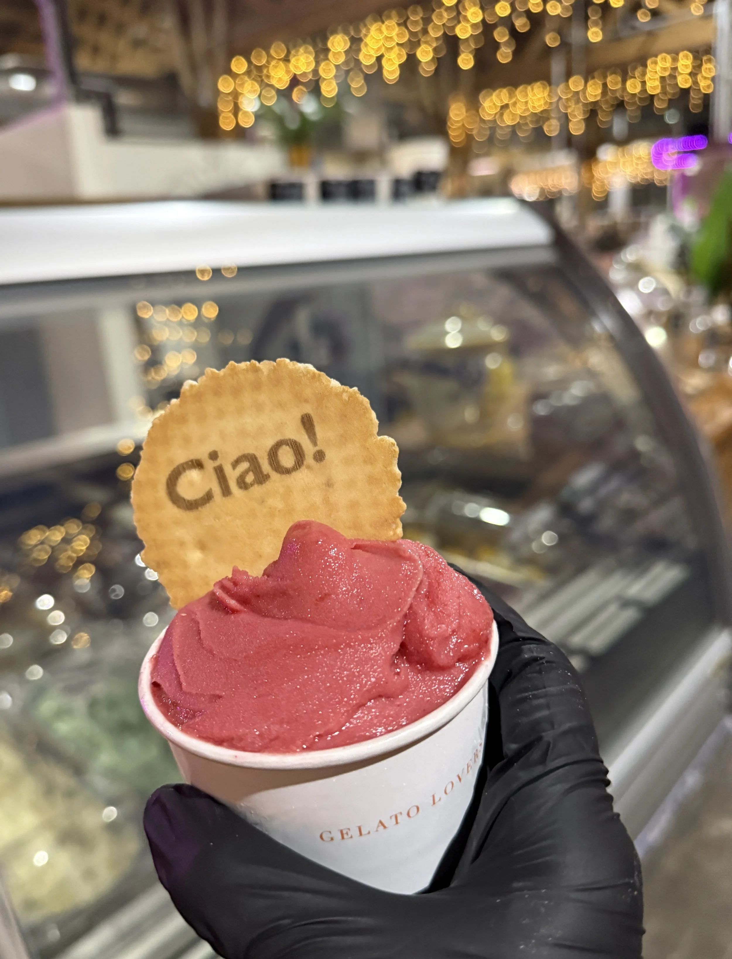 A hand wearing a black glove holding a cup of raspberry sorbet topped with a round waffle cookie that has the word 'Ciao' printed on it, in front of a display case at an ice cream shop with blurred background lighting.