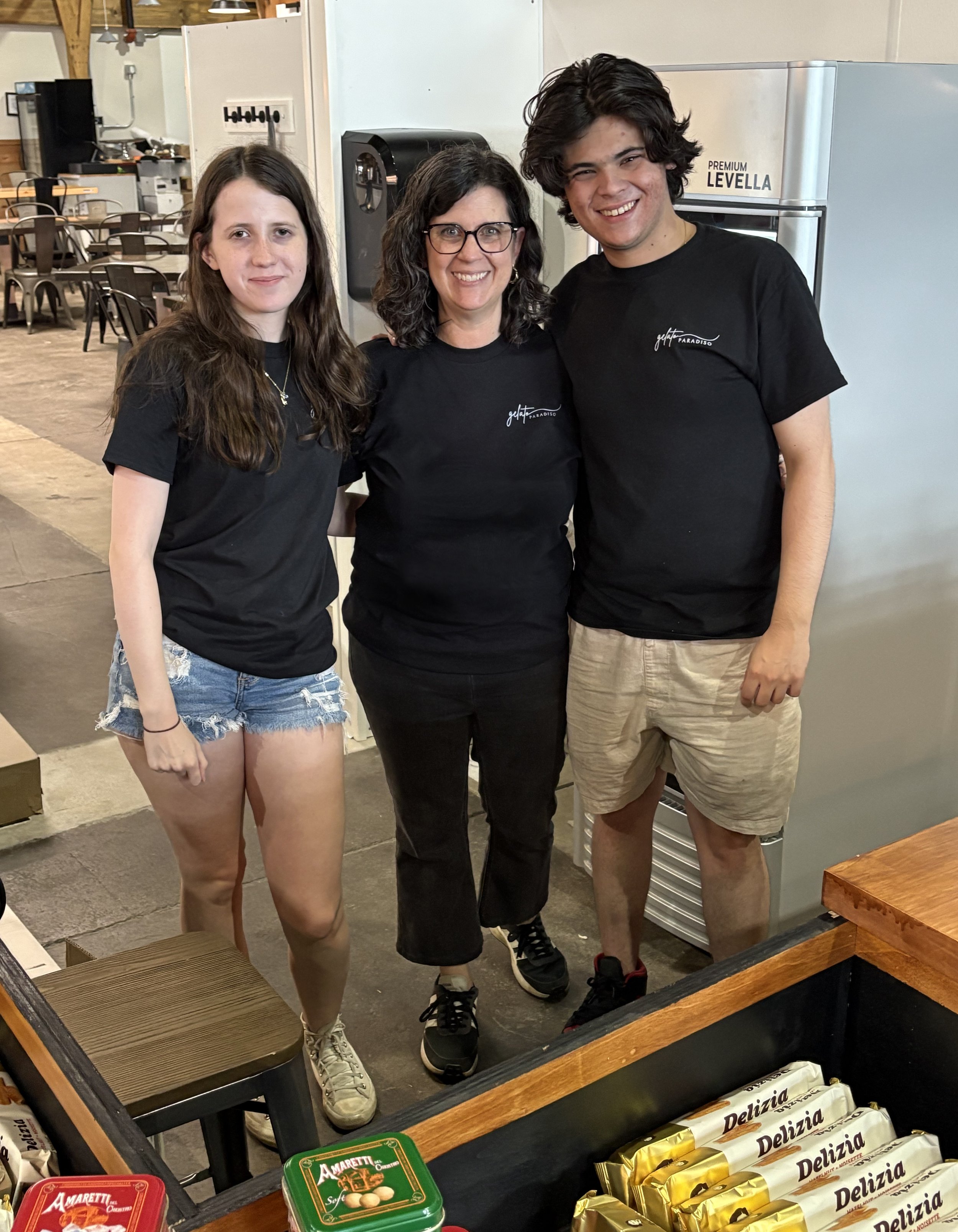 Three people standing inside a store, smiling at the camera. The woman in the middle has curly hair and glasses, the person on the right has dark wavy hair and is wearing a black t-shirt, and the person on the left has long straight hair and is wearing a black t-shirt and denim shorts.