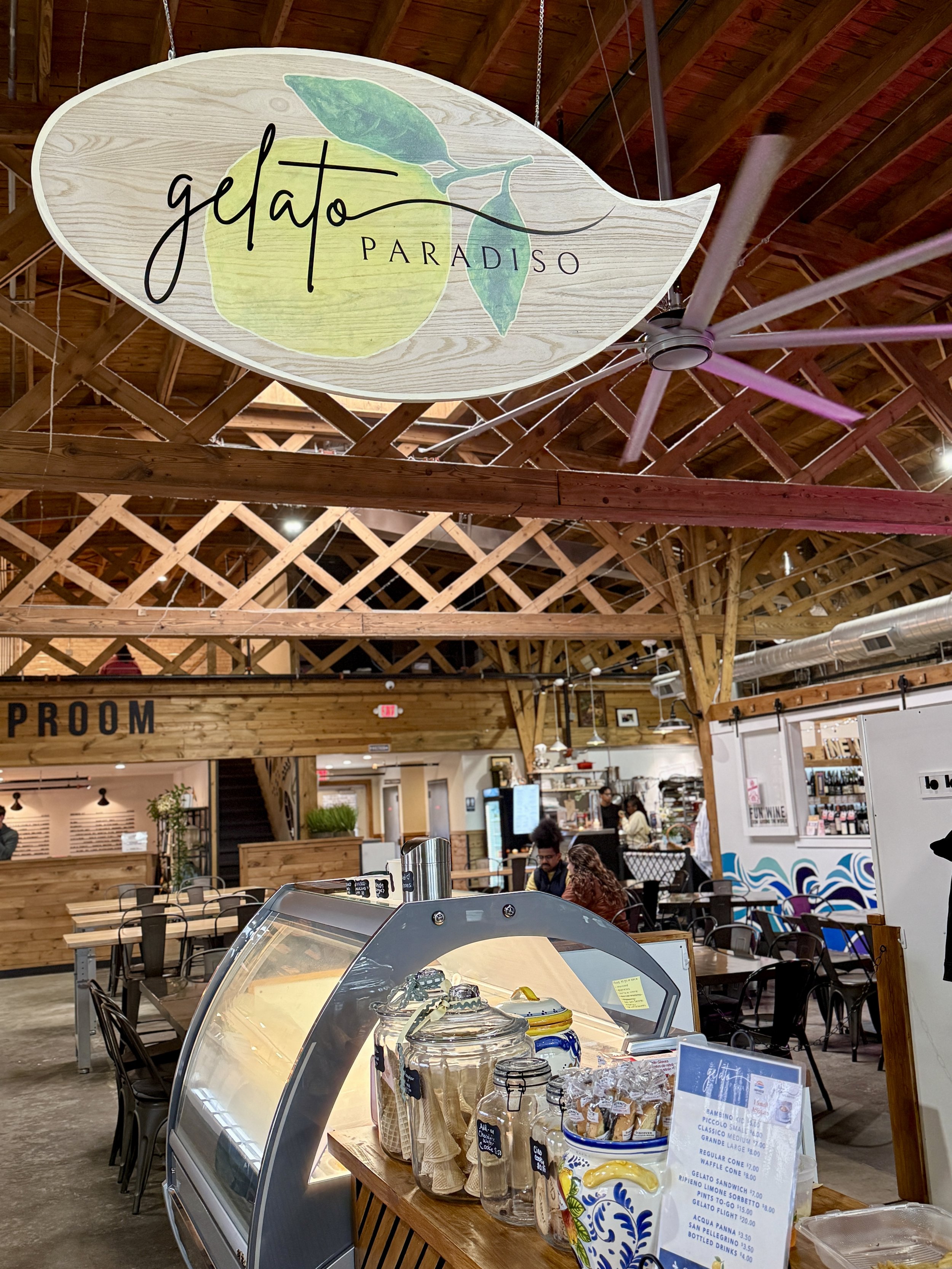 View inside Gelato Paradiso ice cream shop with wooden ceiling, people sitting at tables, an ice cream display case, and a sign hanging from ceiling with the shop's logo featuring a lemon and leaves.