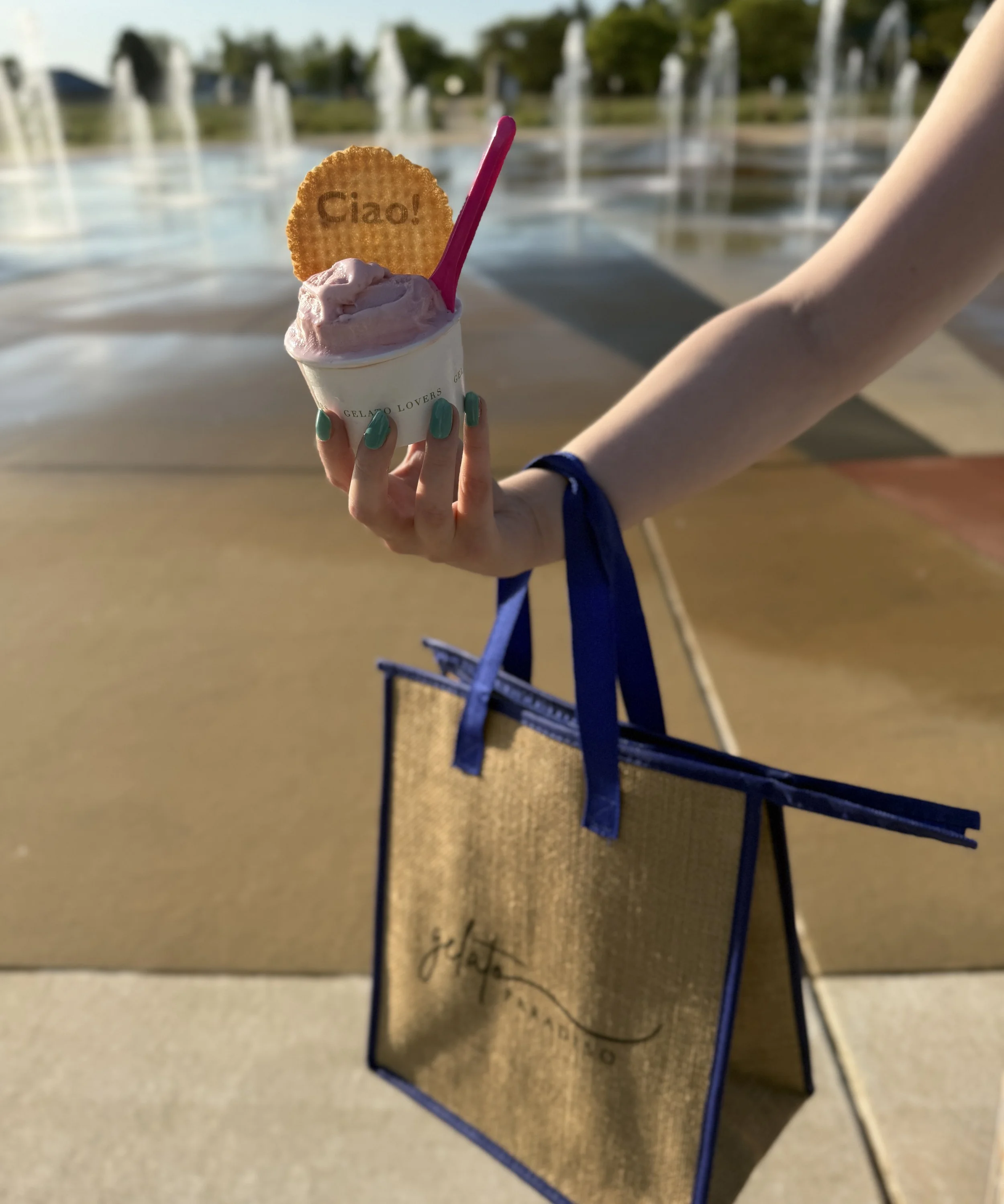 A person's hand holding a cup of strawberry ice cream with a pink spoon and a round cookie that has 'Ciao!' written on it. The background features a fountain with water jets, outdoor trees, and a walkway.
