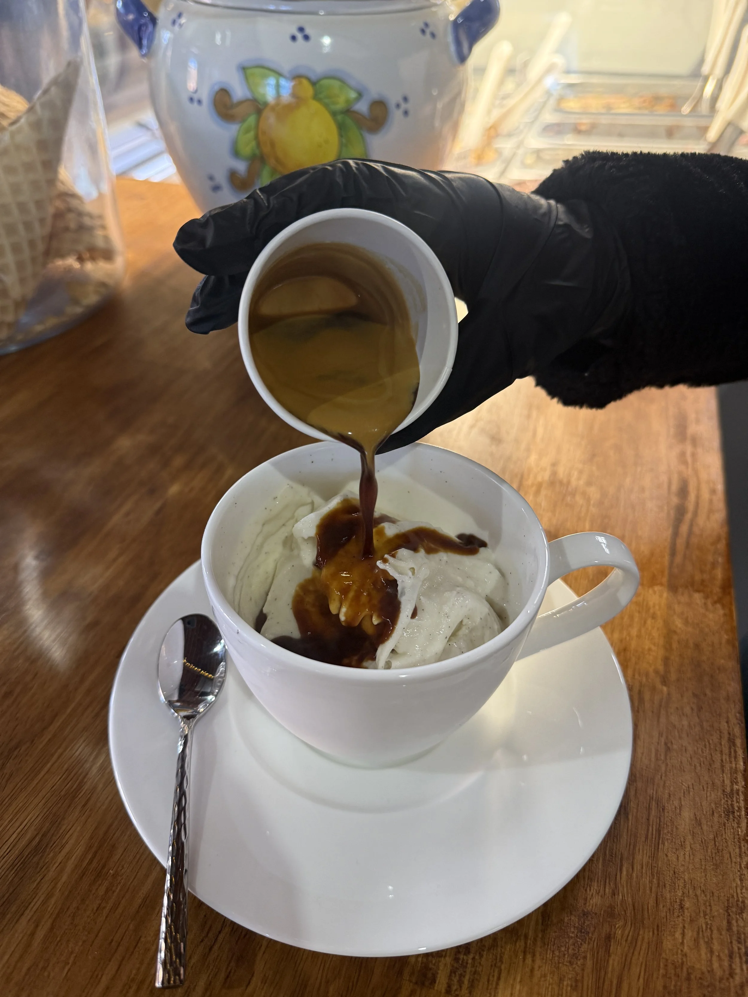 A hand wearing a black glove is pouring espresso over gelato in a mug for an affogato.