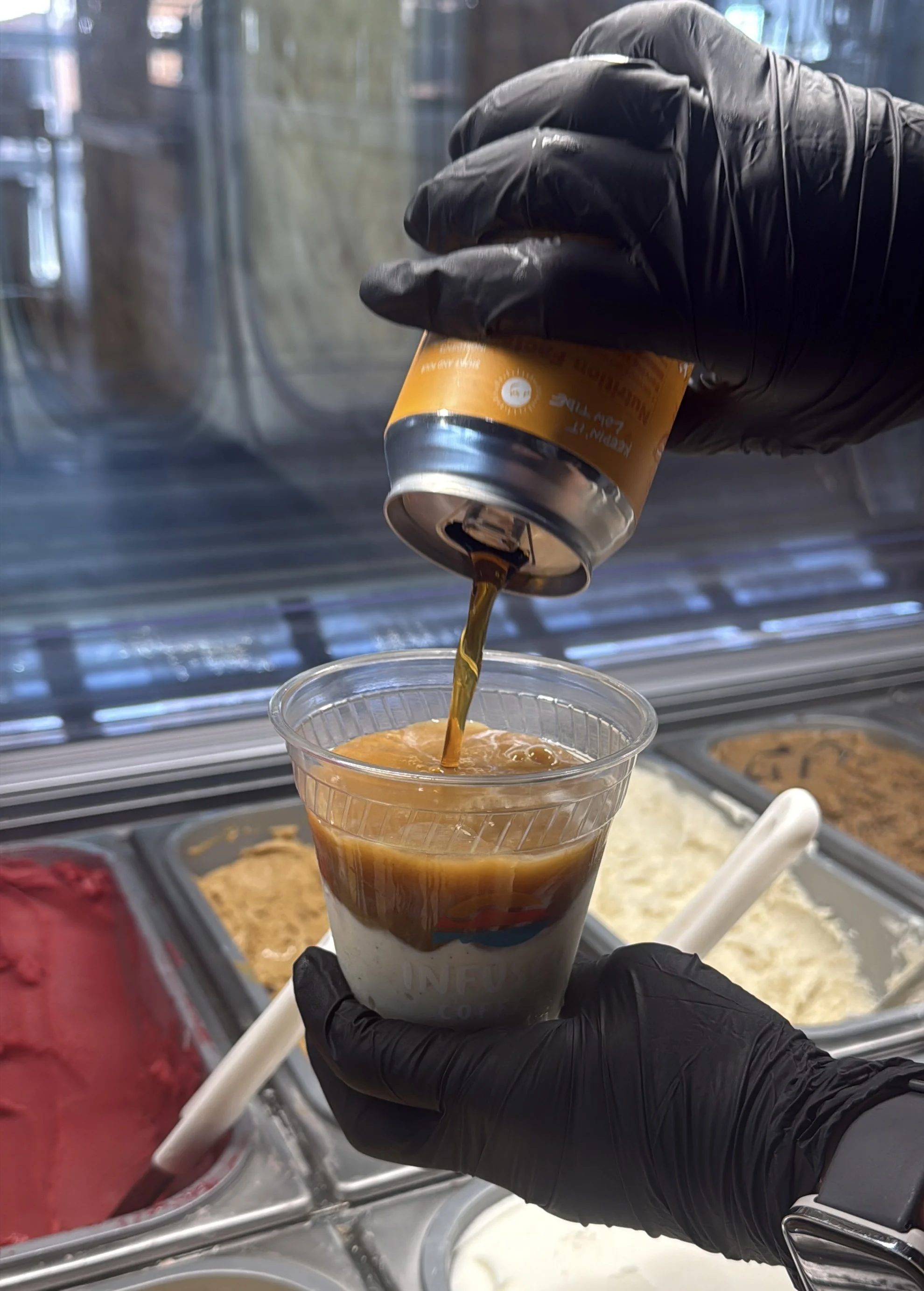 Close-up of a person wearing black gloves pouring cold brew coffee into a clear plastic cup at an ice cream shop or café. In the background, there is a display case with various flavors of ice cream, gelato and sorbetto.