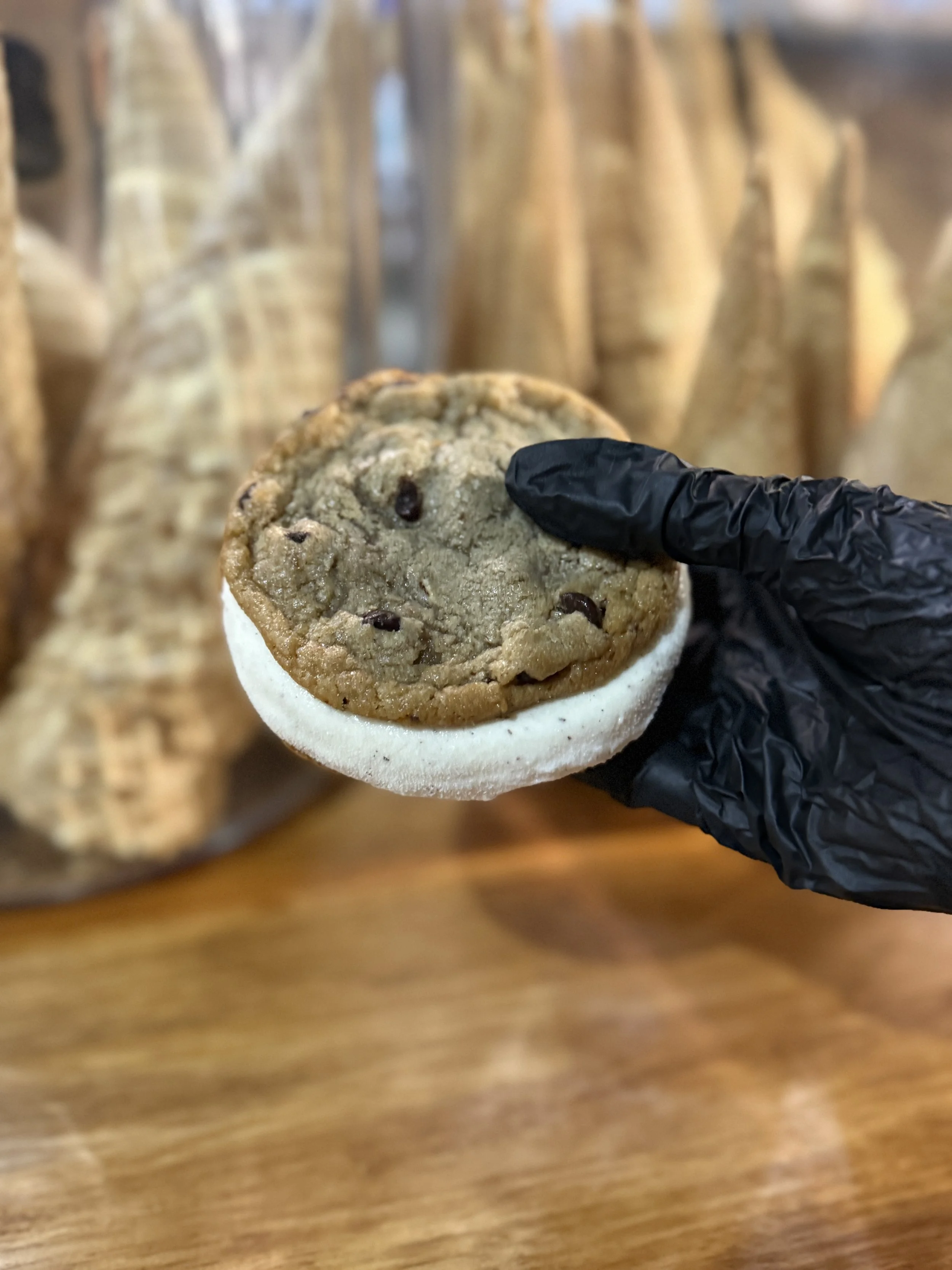 Chocolate chip cookie gelato sandwich held in front of a display of cones in a gelato shop.