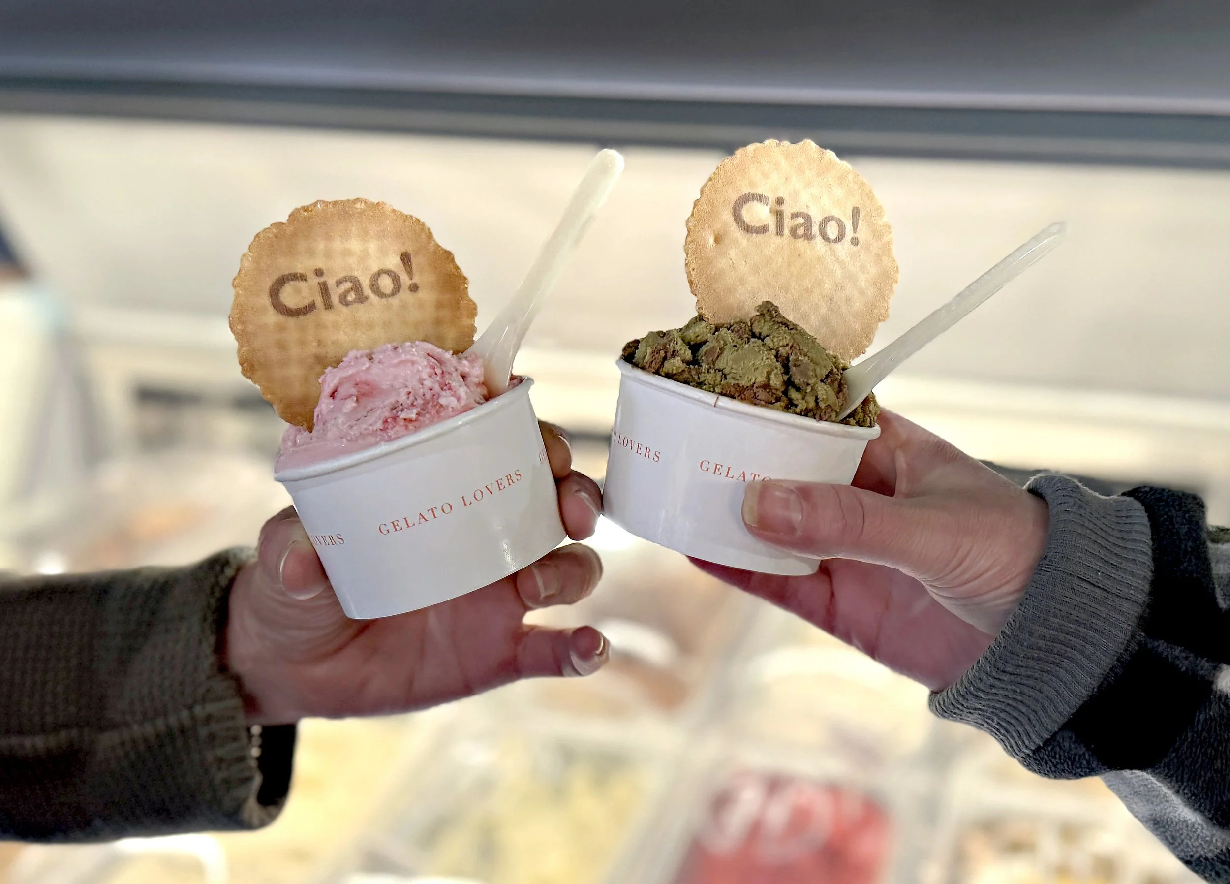 Two cups of gelato, one pink and one green, each topped with a round cookie that has 'Ciao!' written on it, held by two people wearing dark and gray sweaters.