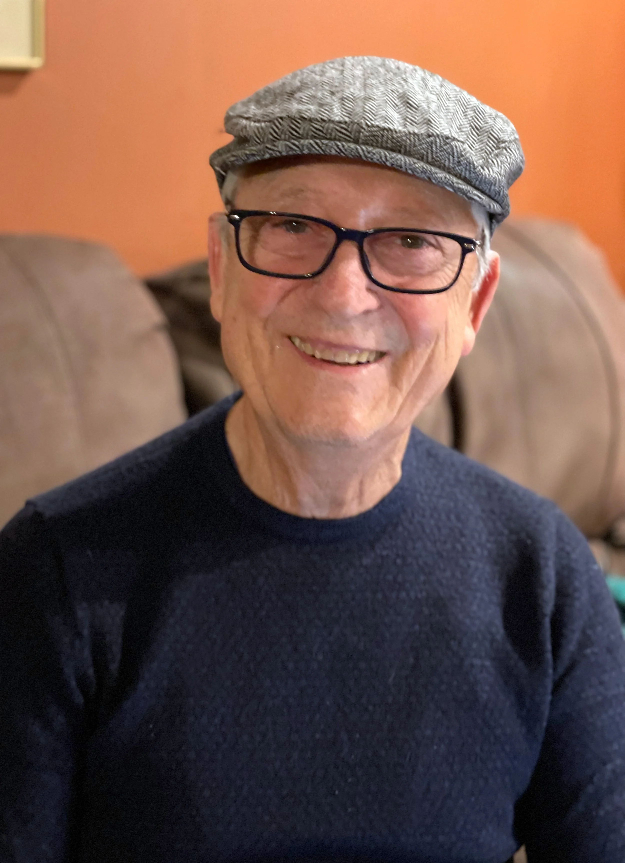 A smiling older man wearing a gray beret, black glasses, and a dark-colored sweater, sitting on a beige couch with an orange wall in the background.