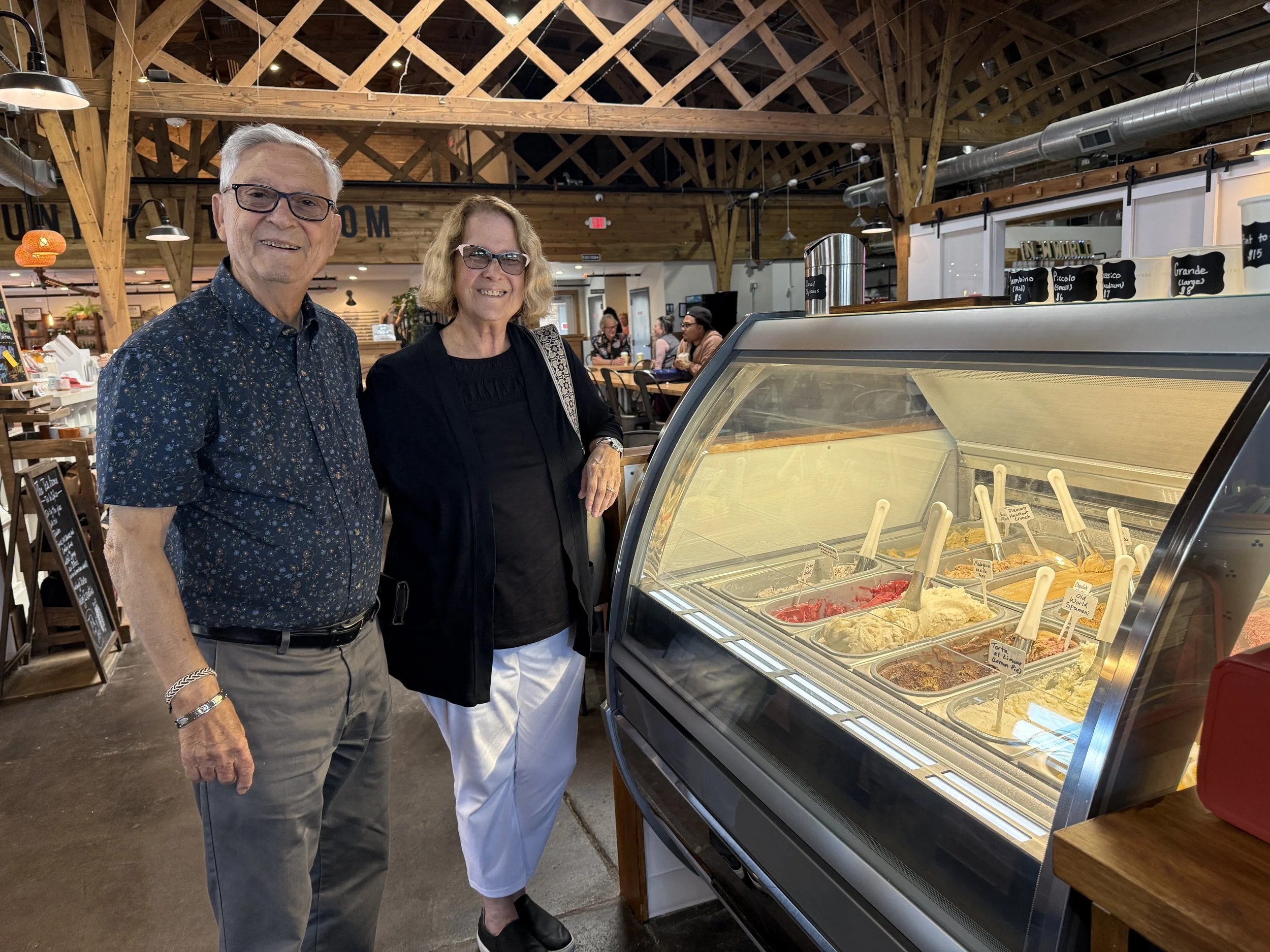 Two smiling older adults, a man with gray hair and glasses and a woman with blonde hair and glasses, stand in front of a gelato display case inside a cafe or ice cream shop with wooden beams and decor.