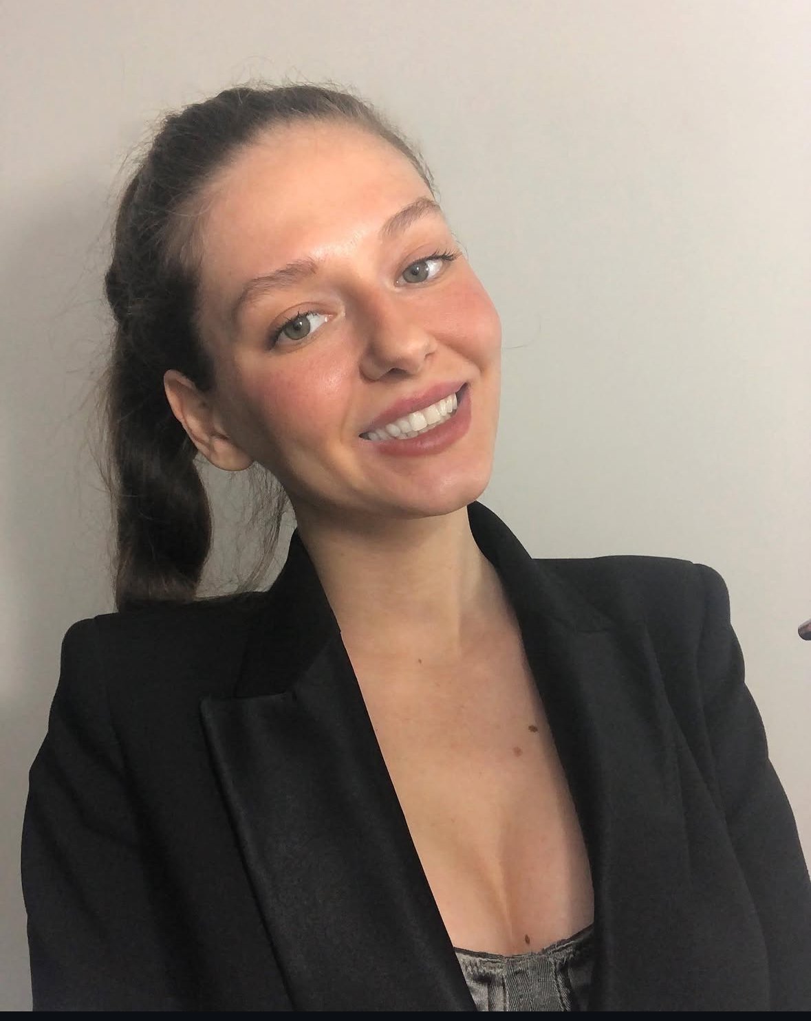 A young woman with light skin, blue eyes, long brown hair tied back, wearing a black blazer and smiling, standing against a plain white wall.