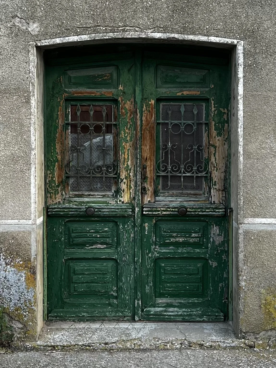 Old, weathered green wooden double door with ornate black ironwork and peeling paint, set in a gray concrete wall.