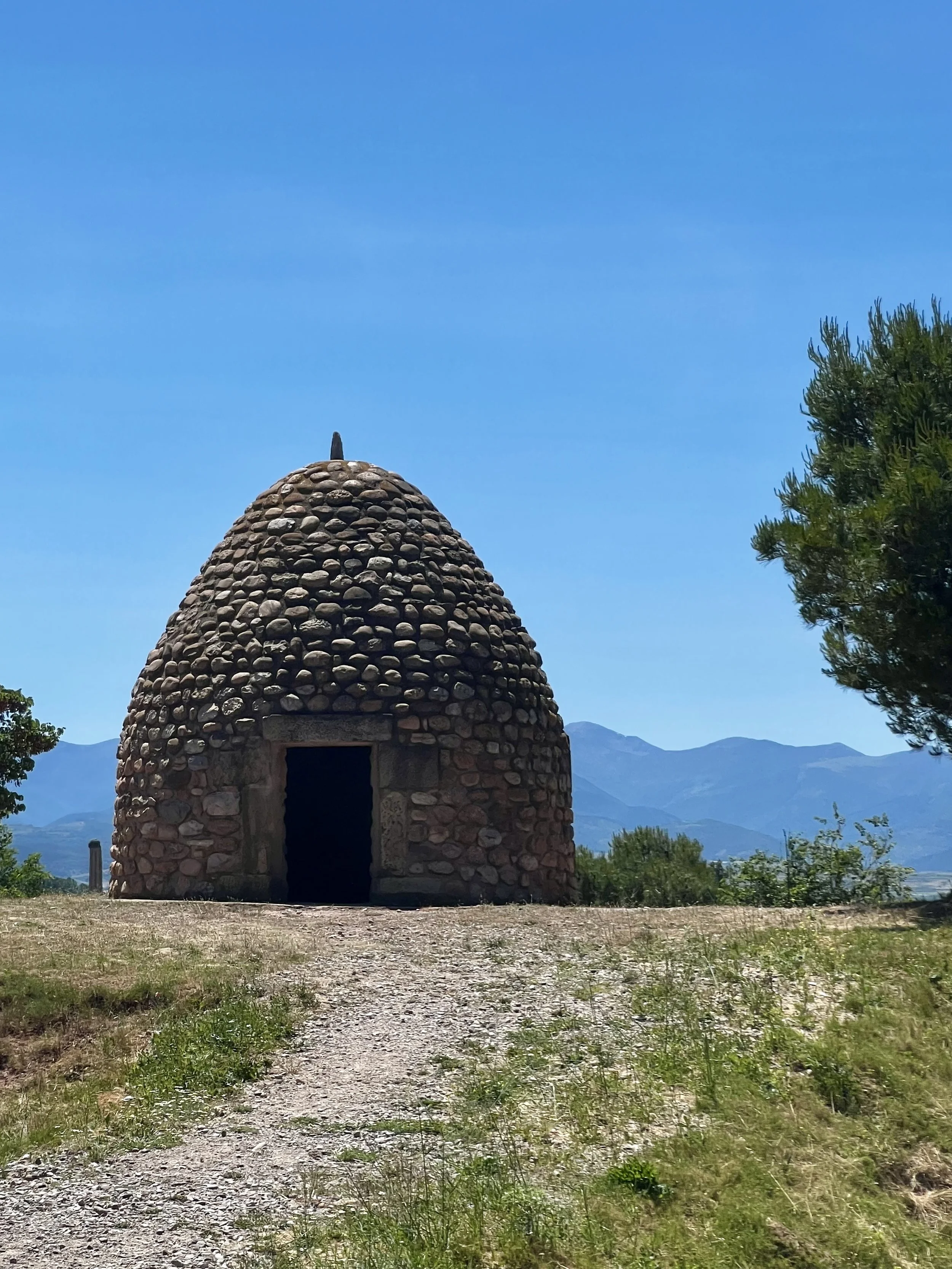 A small round stone building with a conical roof and dark doorway, set in a sunny outdoor landscape with mountains in the background and clear blue sky overhead.