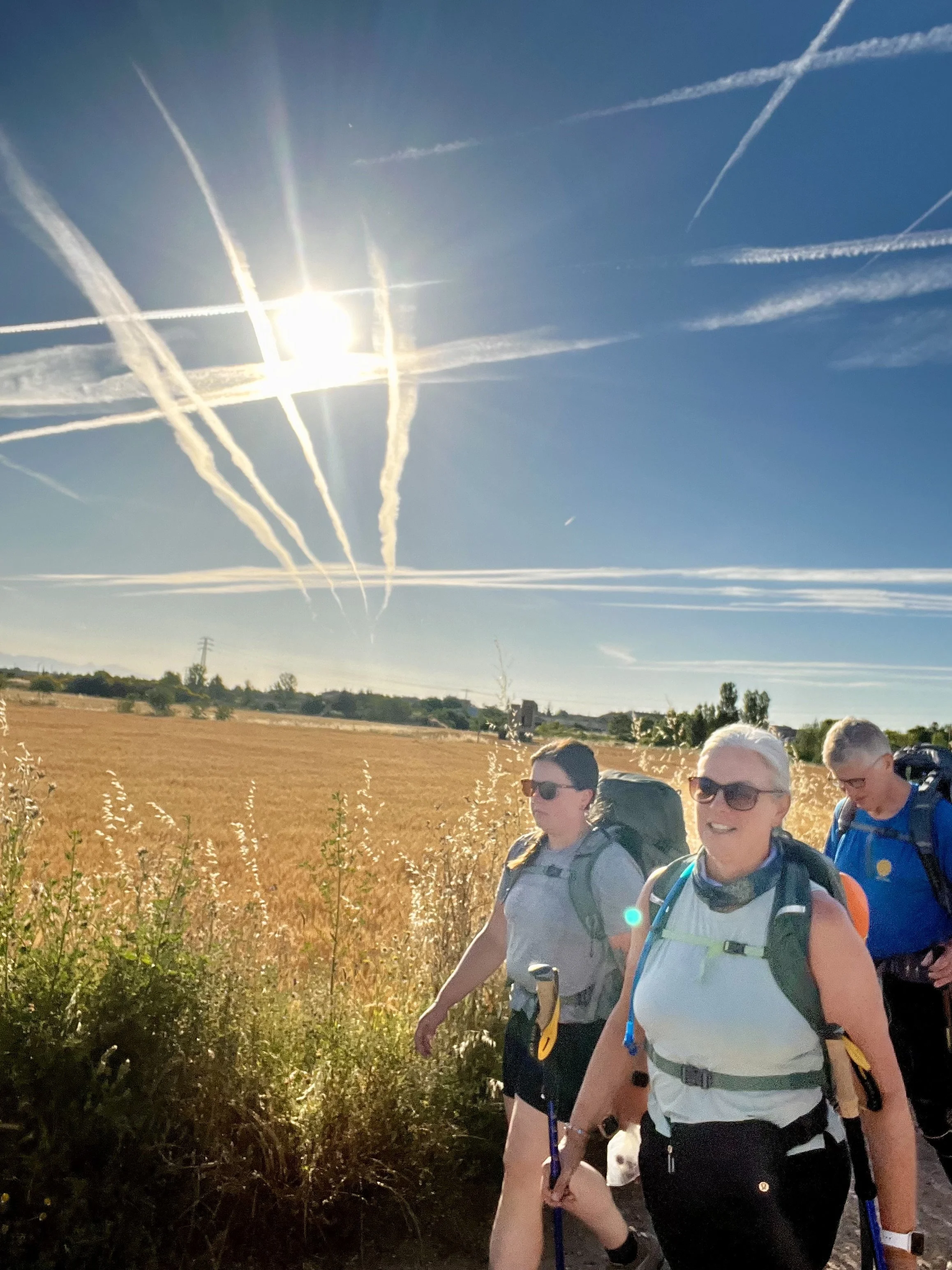 Four hikers walking along a trail with a rural landscape, including fields and trees, under a bright sun in a clear blue sky with contrails.