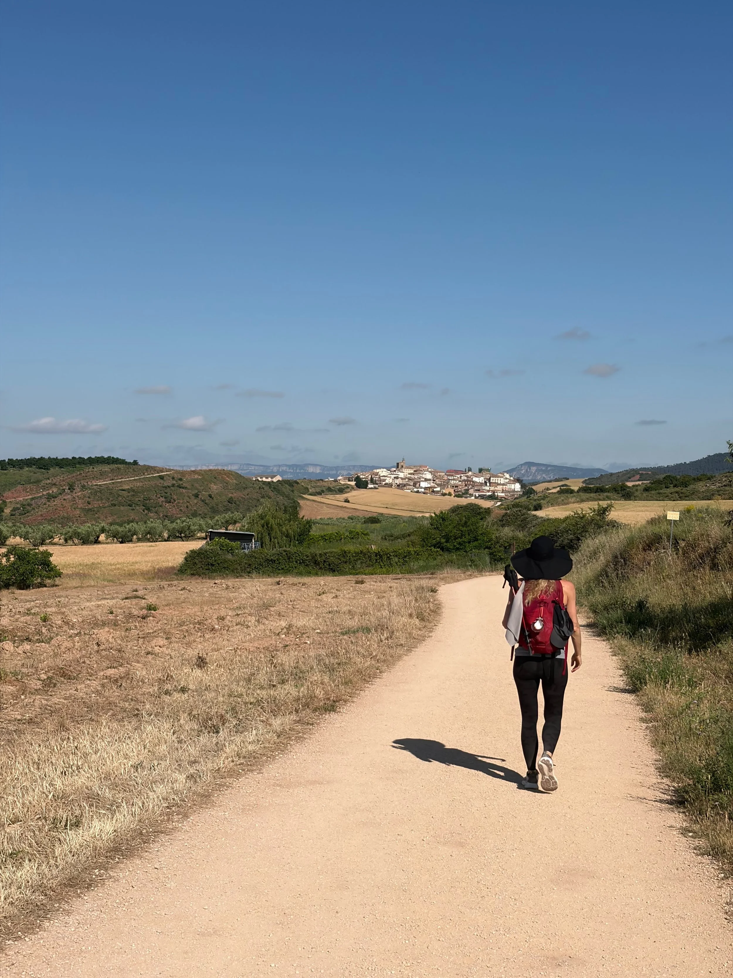 A woman wearing a black hat, red backpack, and black pants is walking along a dirt path in a rural area with fields and greenery, with a small town visible in the distance under a blue sky with some clouds.