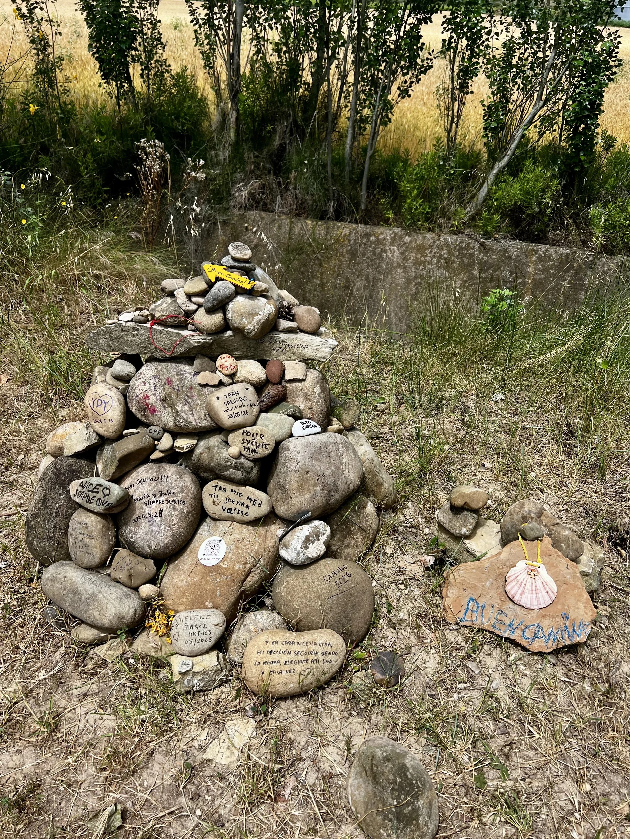 A small memorial altar made of stacked rocks with handwritten messages on some stones, in front of a grassy and wooded area.