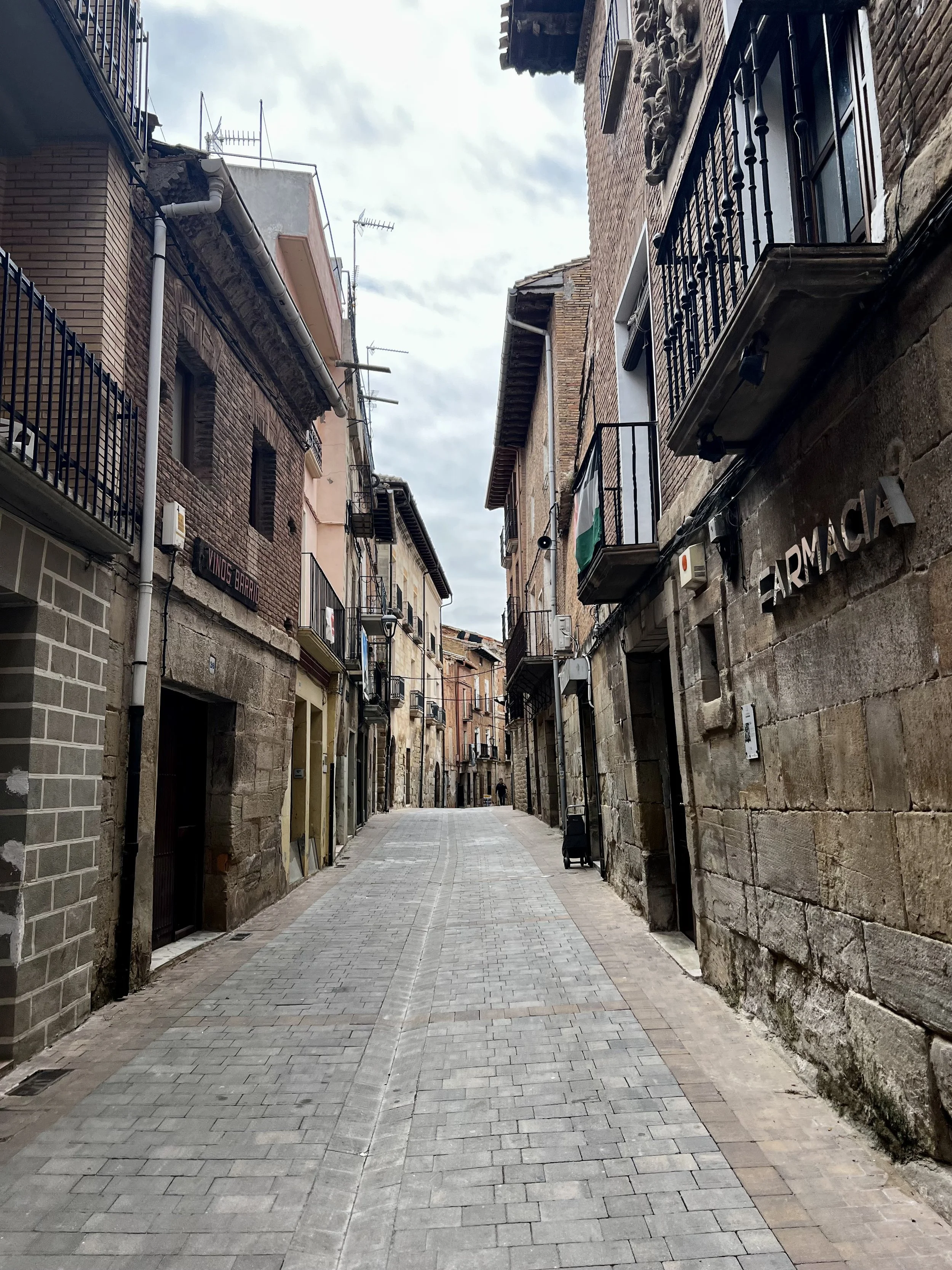 Empty cobblestone street lined with old brick buildings on both sides, some with balconies and signage, under a cloudy sky.