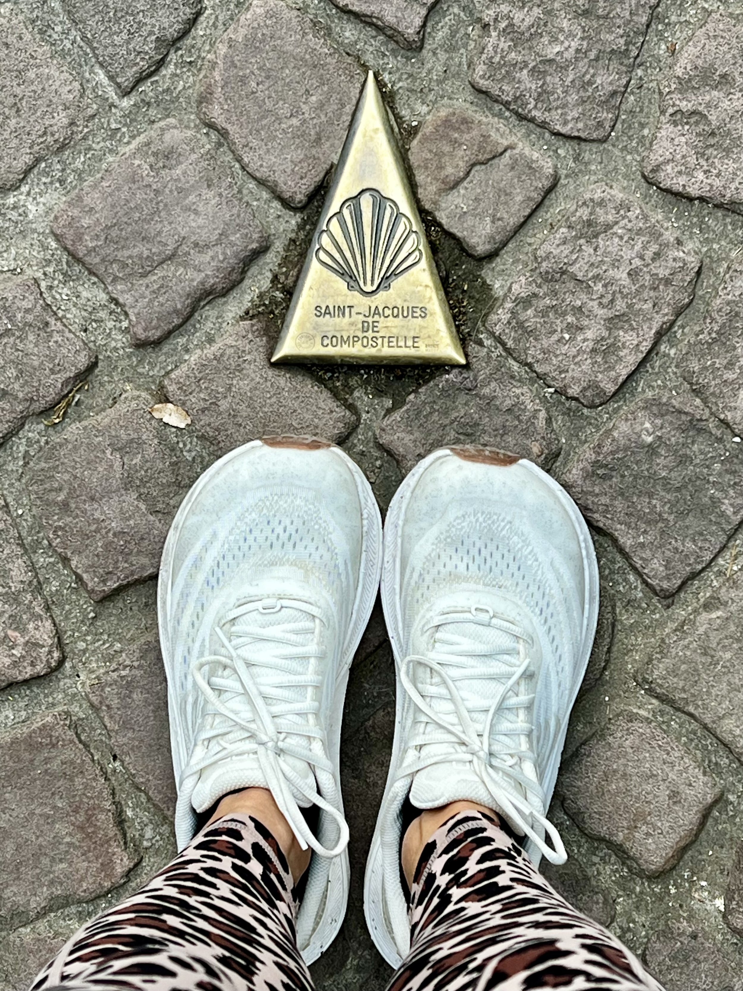 A pair of white sneakers on a cobblestone street, with a gold triangular marker labeled 'Saint-Jacques de Compostelle' in front of them, and a person wearing animal print leggings.