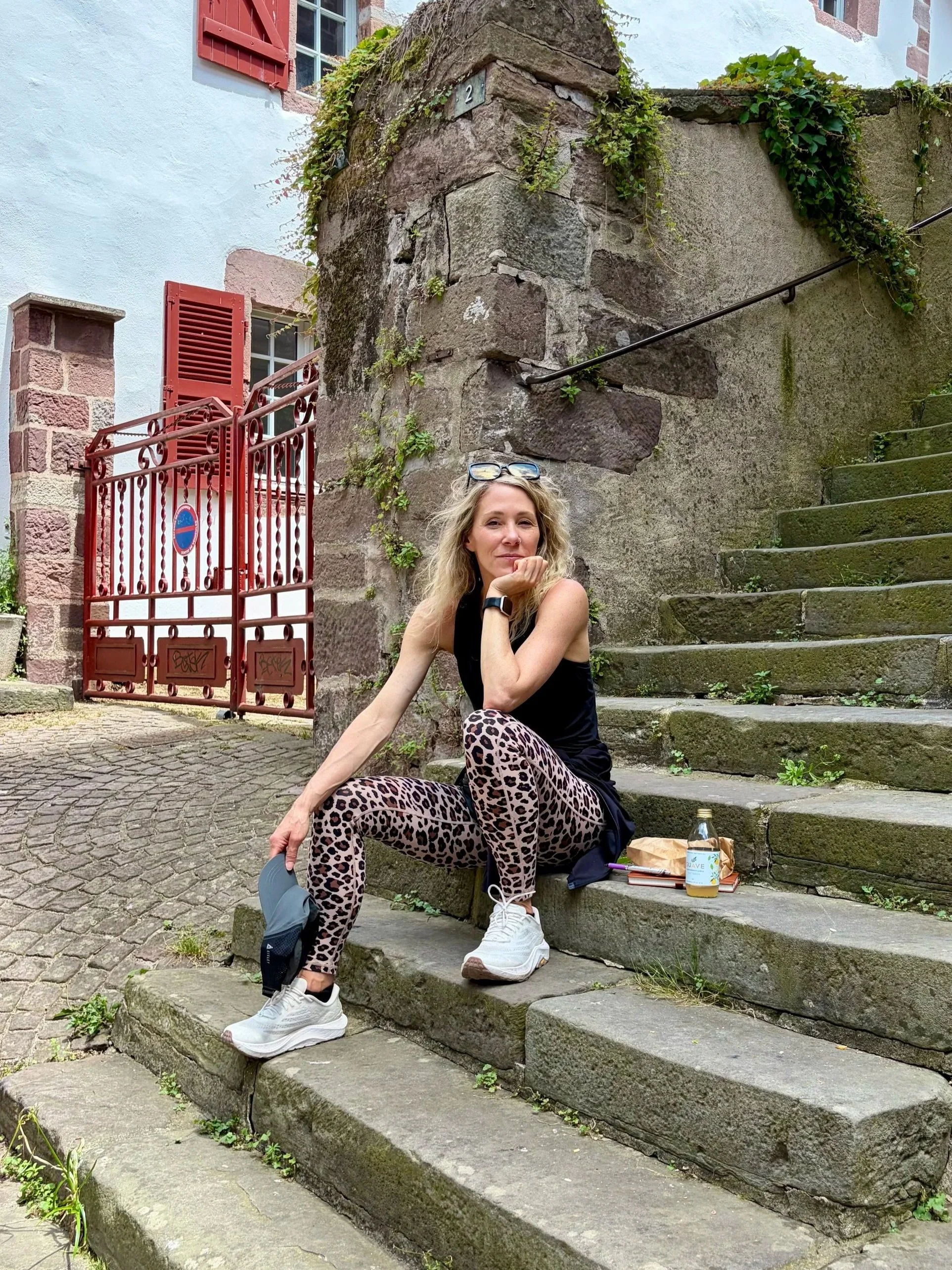 A woman sitting on stone stairs near a rustic stone wall with plants growing on it. She has curly blonde hair, is resting her chin on her hand, and is wearing leopard print pants, a black sleeveless top, and white sneakers. She has glasses on her head, a smartwatch on her wrist, and there are a water bottle and some personal items placed on the stairs beside her.