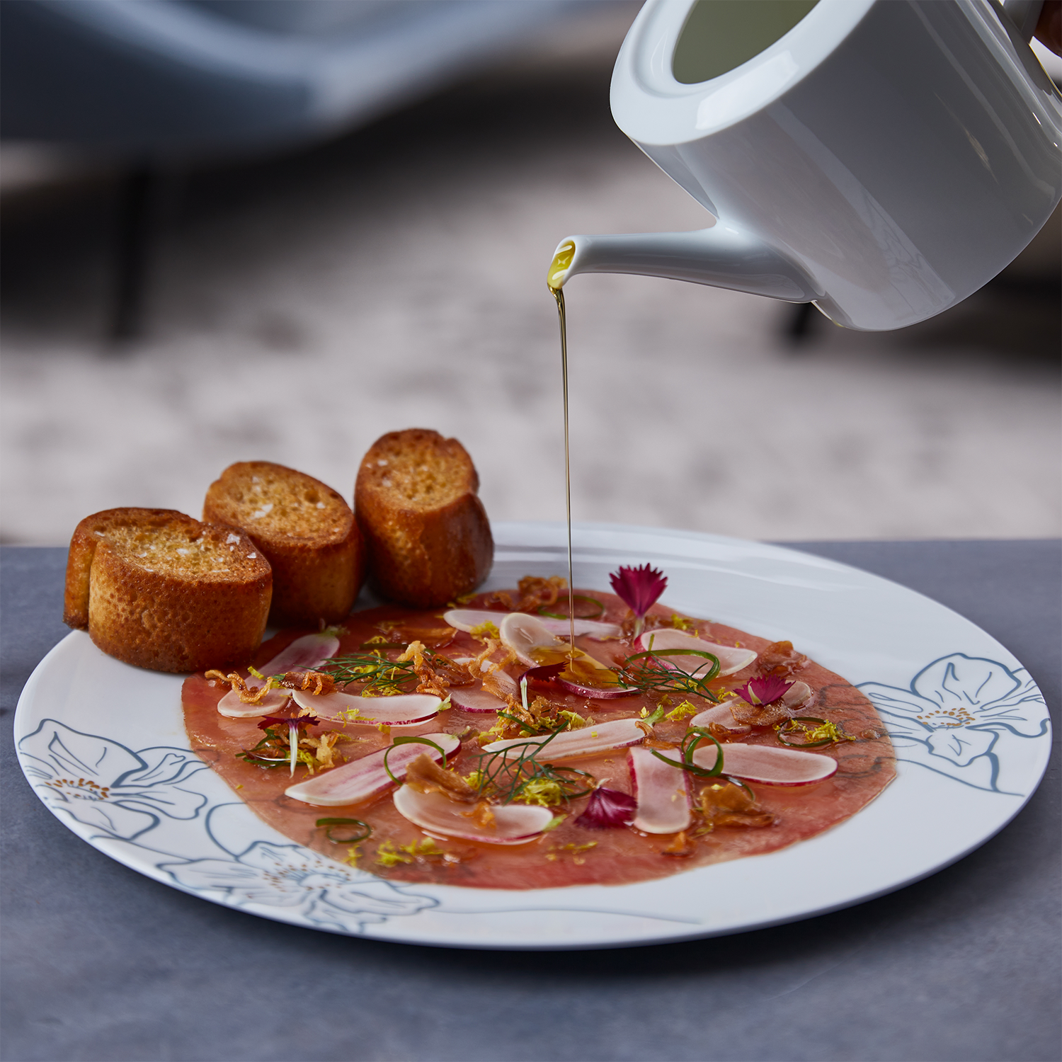 A plate of carpaccio with sliced meat, garnished with herbs, onion slices, and edible flowers, with toasted bread on the side. Olive oil is being drizzled on the dish.