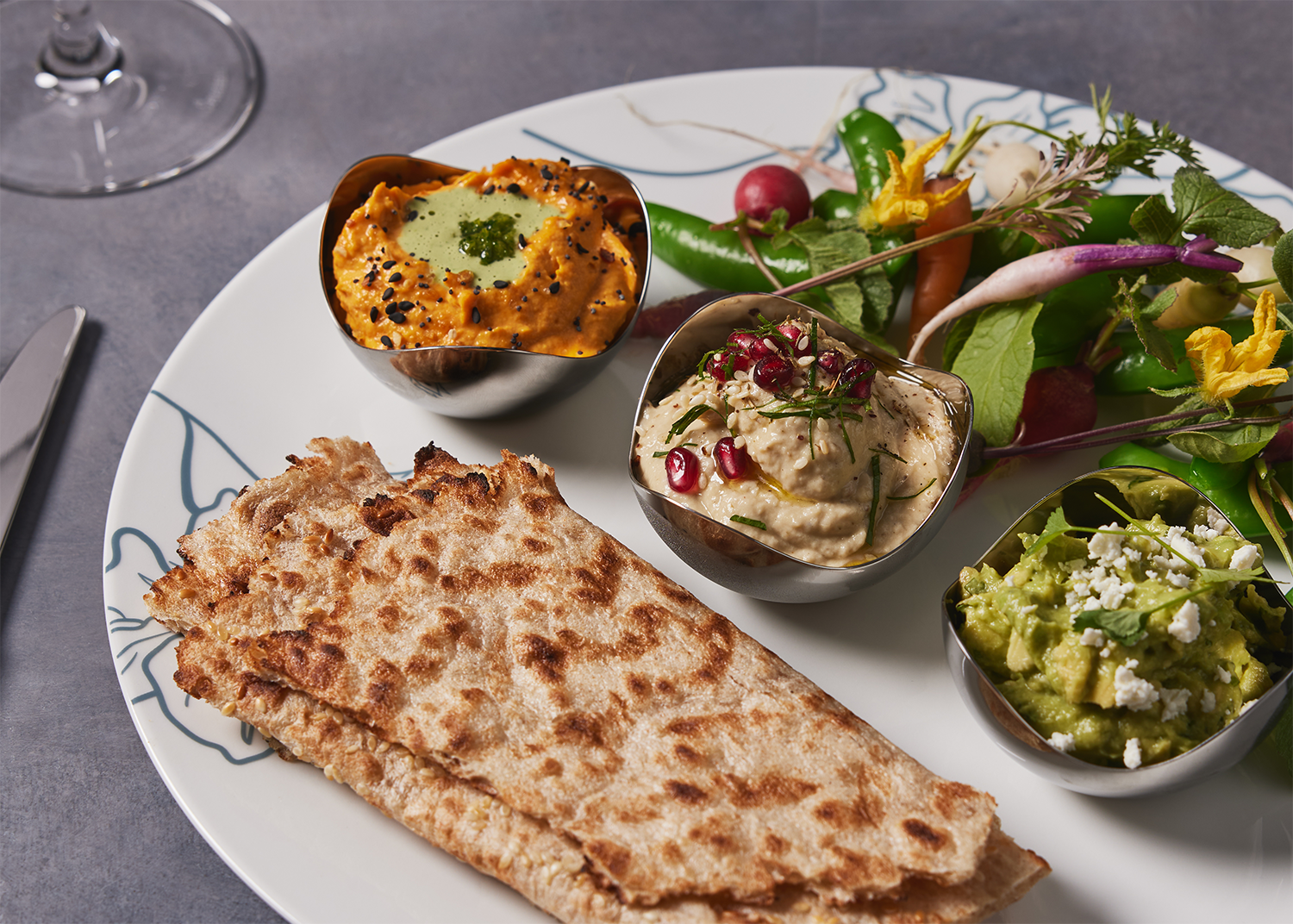 Platter dip featuring naan bread, three bowls of dips and condiments, and fresh salad greens with radishes and edible flowers.