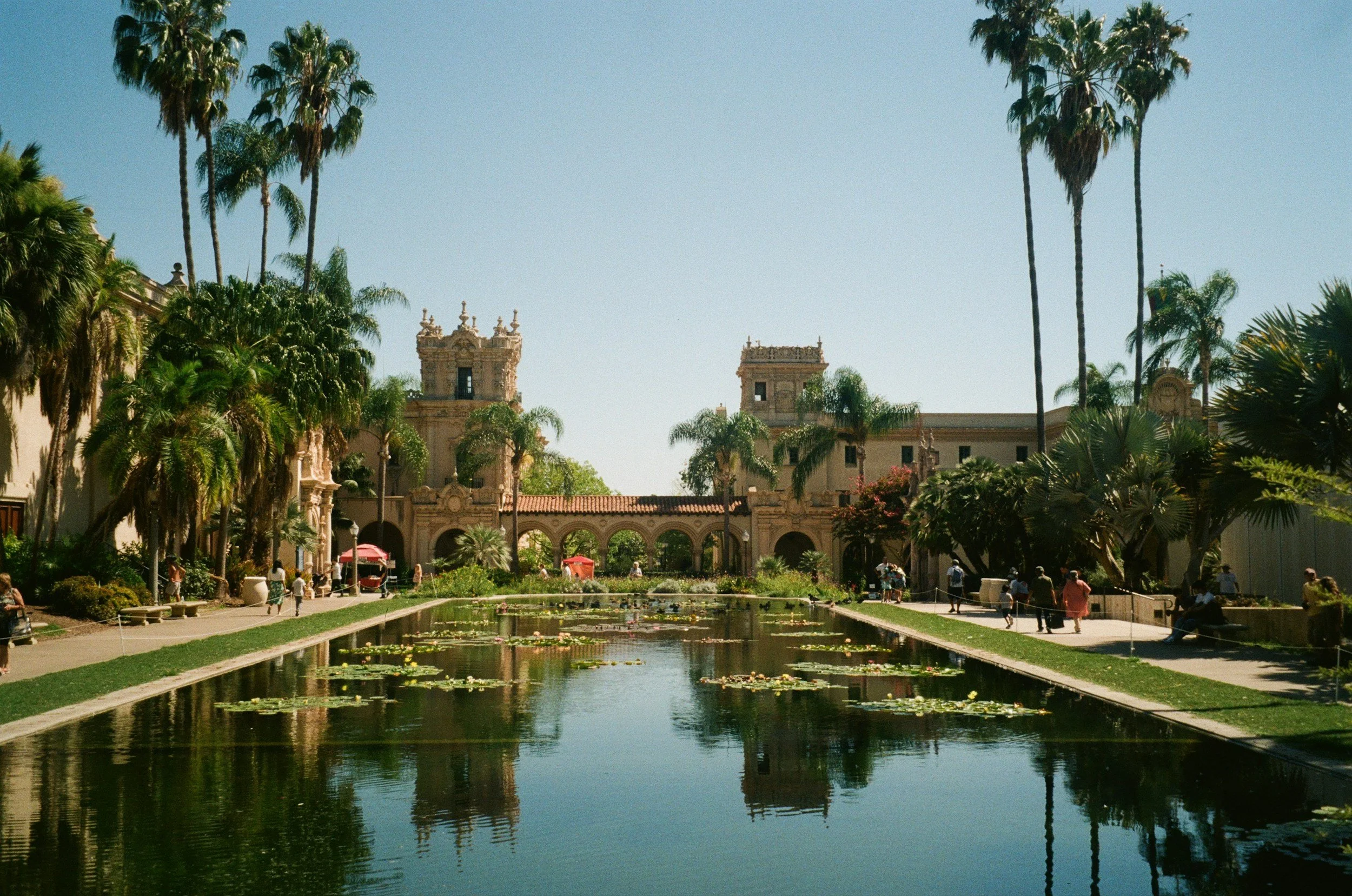 A park with a water feature and lily pads in the foreground, surrounded by trees and historic buildings with towers and arches, sunny day with clear blue sky.