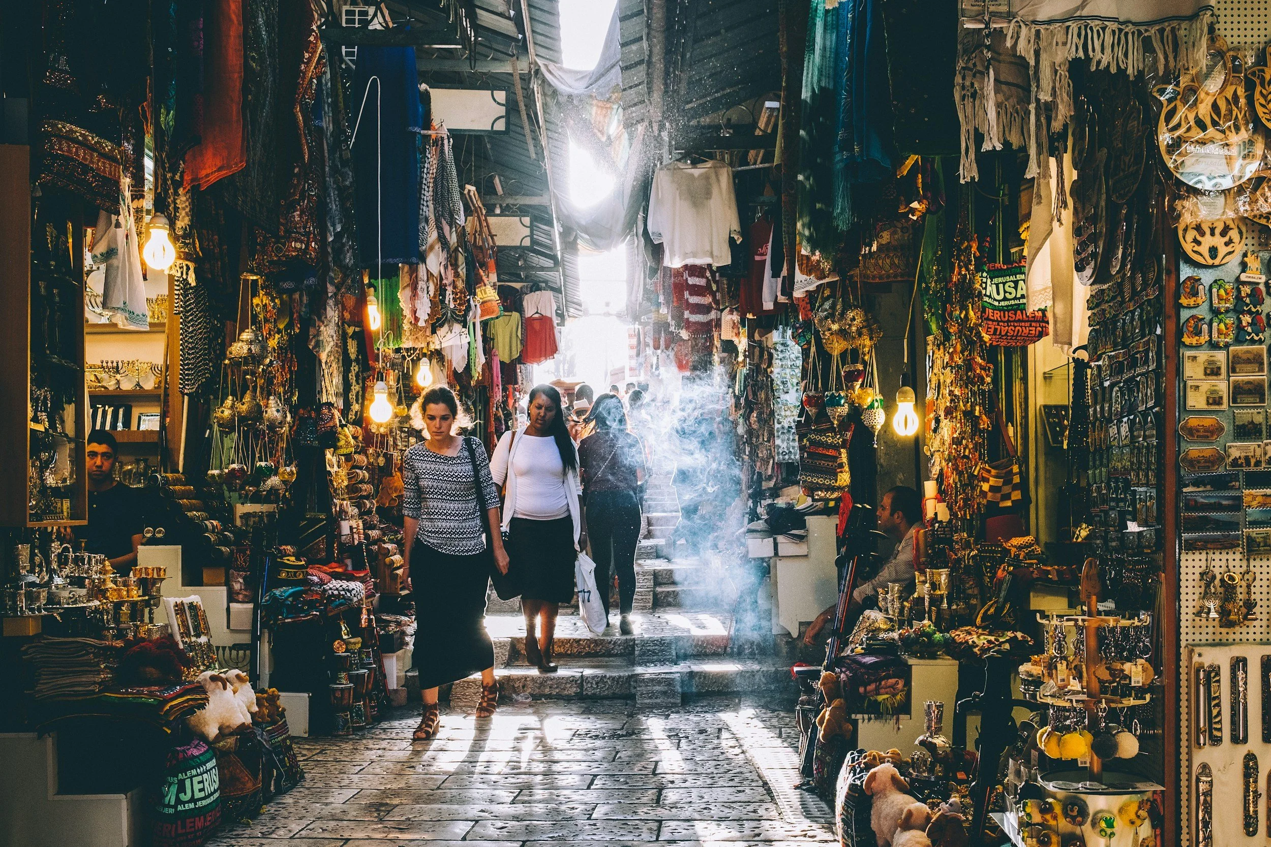 A marketplace with shops displaying jewelry, clothing, and souvenirs. Several shoppers walk through the narrow aisle, with sunlight streaming in from behind, creating shadows on the cobblestone floor. The shops are decorated with colorful items and hanging textiles.