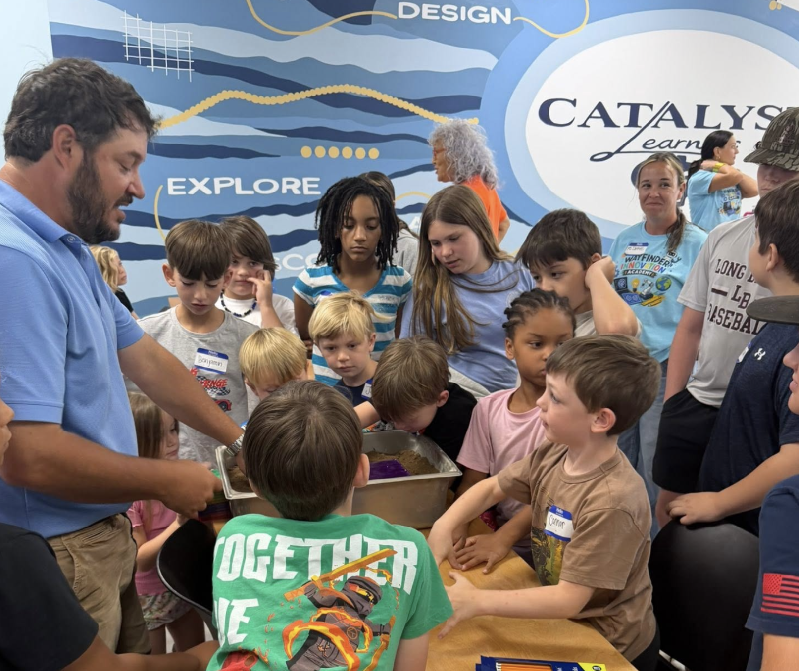 Children and adults gathered around a table at an event, with a man in a blue shirt demonstrating something. The background has a mural with words like 'Design,' 'Explore,' and 'Catalyst Learning'.