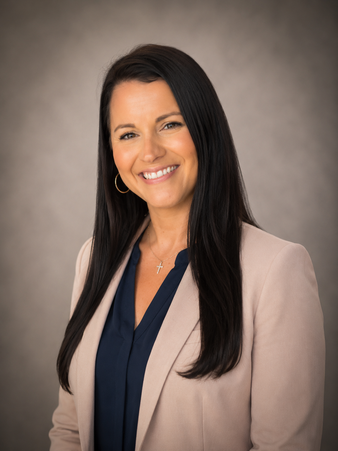 Professional headshot of a smiling woman with long dark hair, wearing a beige blazer, navy blouse, gold hoop earrings, and a cross necklace against a neutral background.