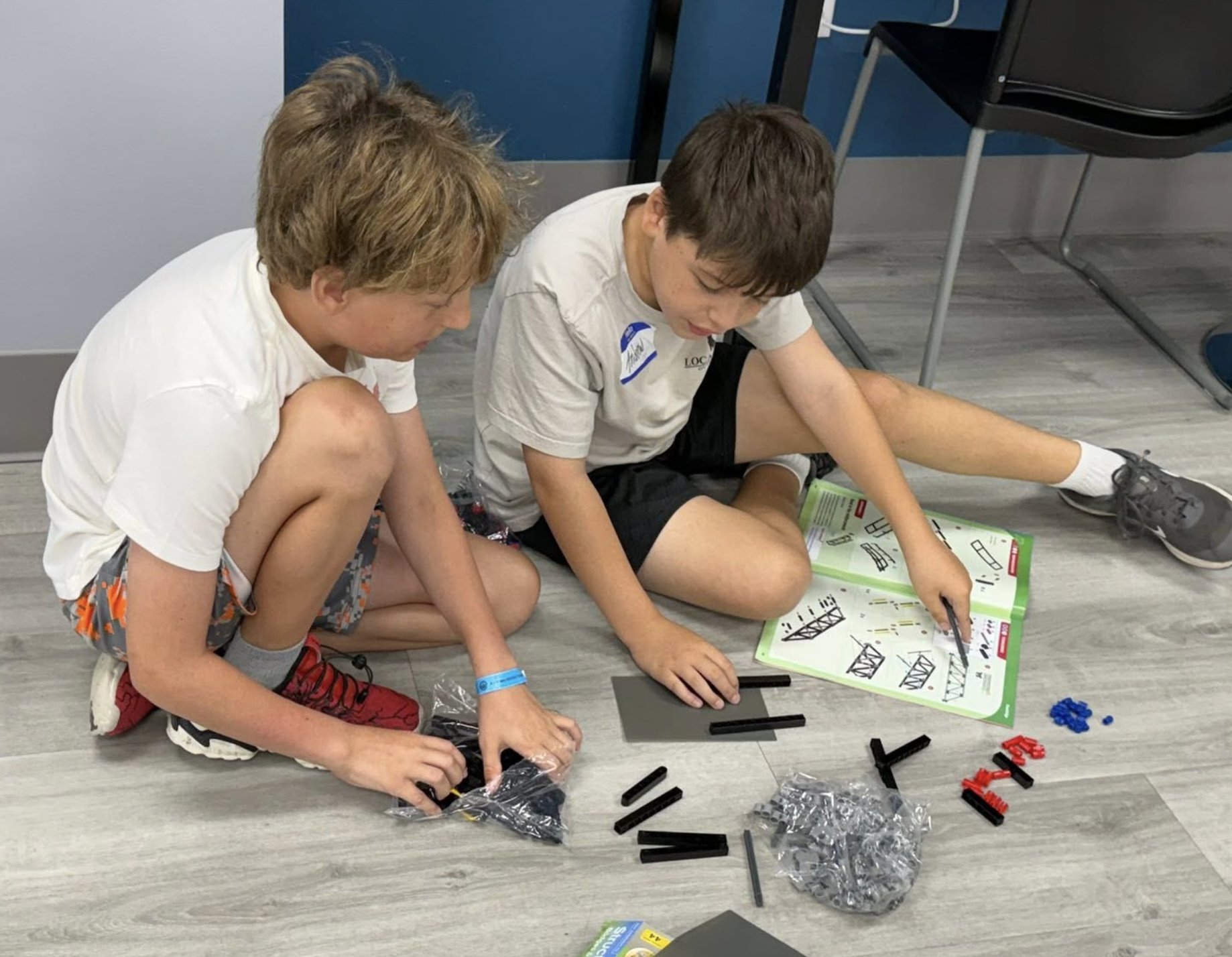 Two boys sitting on the floor assembling pieces of a plastic building set, with an instruction sheet showing bridge diagrams nearby.