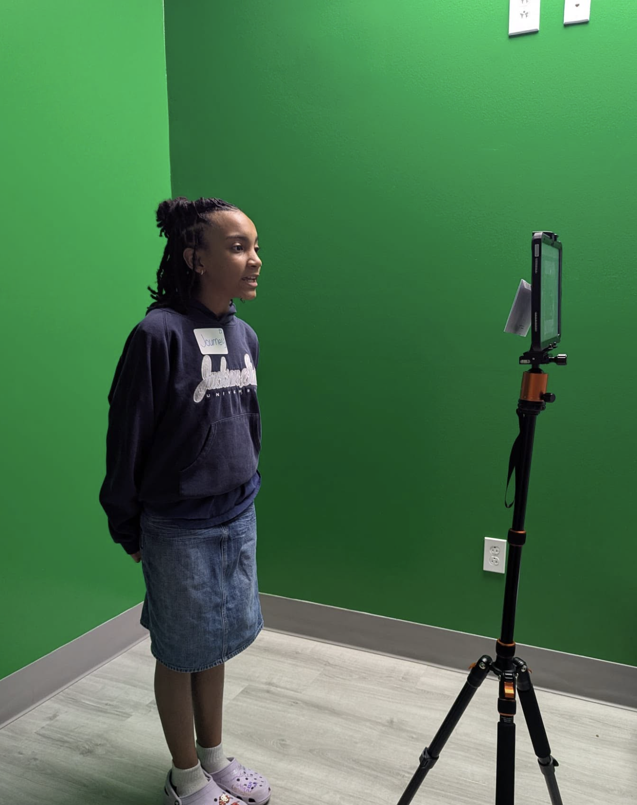 A young girl with braided hair, wearing a navy hoodie, denim skirt, and purple Crocs, standing against a green wall and looking at a tablet on a tripod stand in a room with gray flooring.