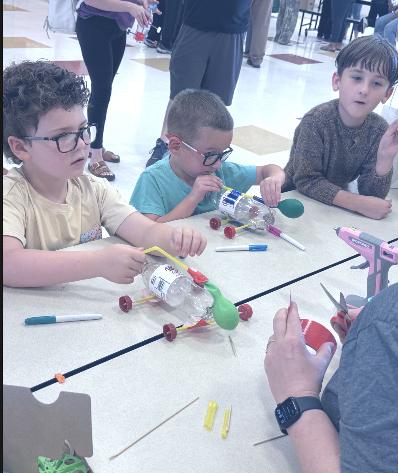 Children sitting at a table building cars with plastic bottles, straws, and craft supplies, with adults in the background.