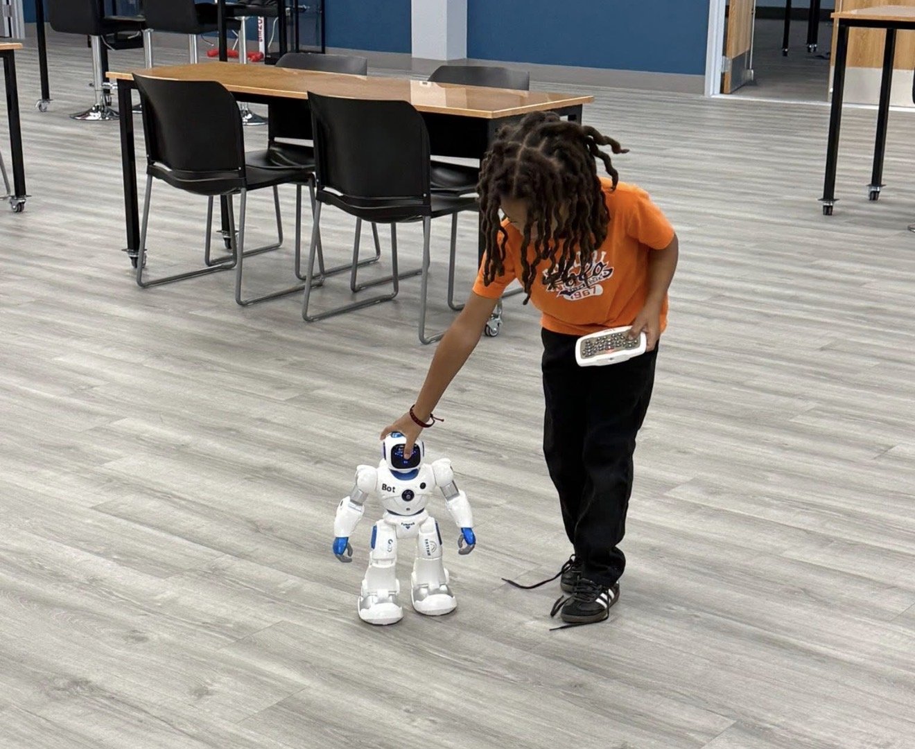 A young person with dreadlocks, wearing an orange T-shirt and black pants, is interacting with a small white robot on a light-colored wooden floor in a modern room with tables and chairs.