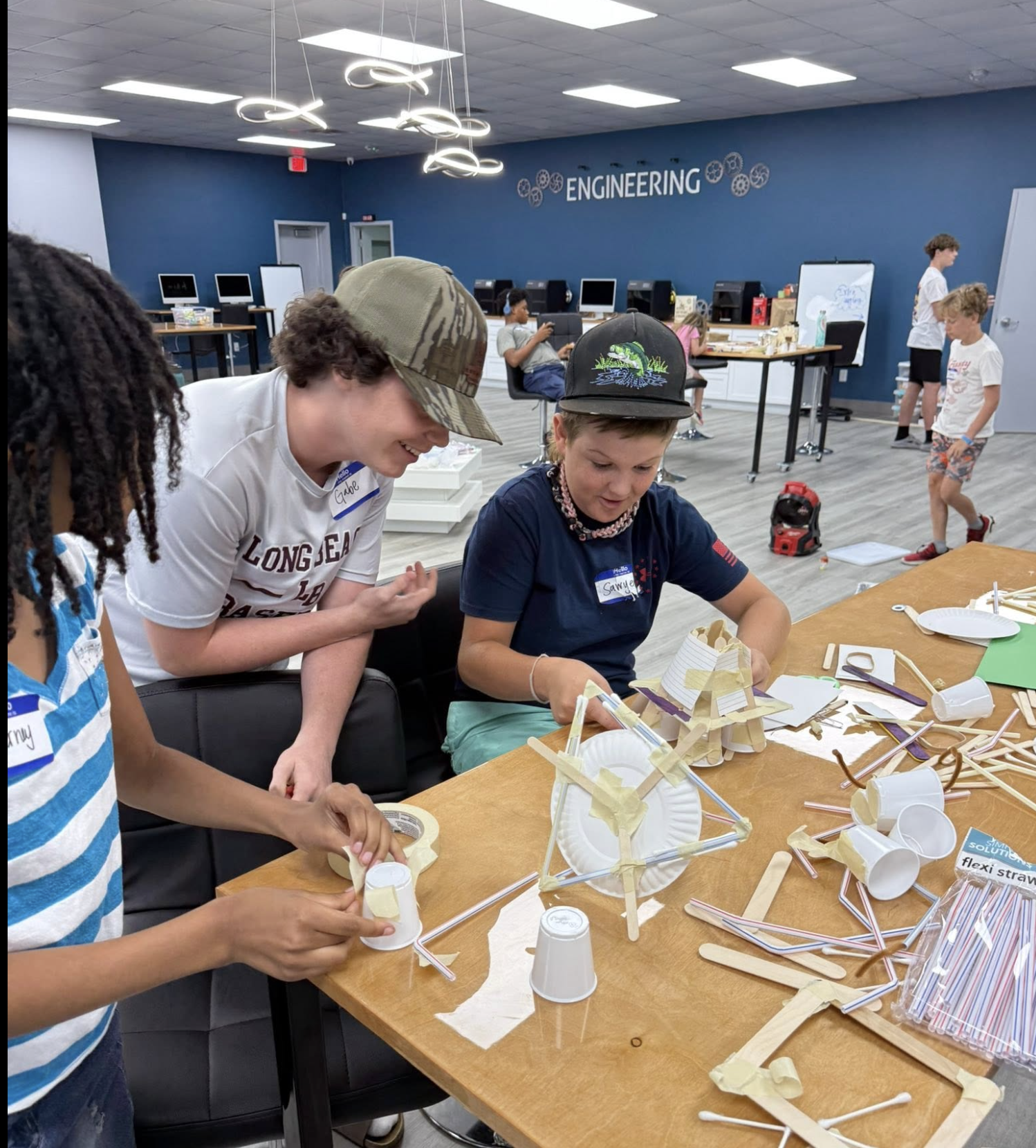 Children and teenagers engaged in a STEM activity in a classroom labeled 'Engineering'. They are working on constructing structures using paper cups, straws, tape, and popsicle sticks. The room has computers against the wall and a blue and white color scheme.
